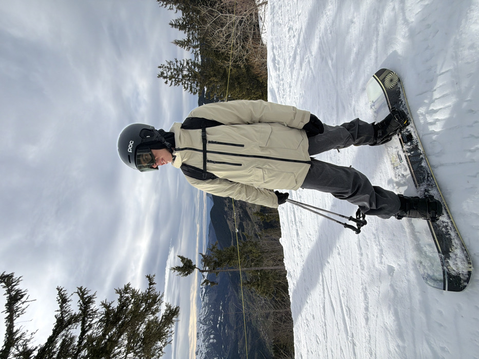An individual is depicted standing on a snow-covered mountain slope in Manigod, France. The person, viewed from the waist up, wears a dark grey POC-branded helmet with dark goggles, a black balaclava, a light beige winter jacket with black zippers and pockets, dark grey snow pants, and black gloves. A black backpack is worn over the jacket. The individual is positioned on a snowboard with black bindings, which features a black and white base with a spiral design near the tail. Two black and silver ski poles are held in the person's left hand. The individual is looking towards the right side of the frame. The immediate environment consists of groomed snow. In the background, a coniferous tree is visible on the left, and several more scattered pine trees are present across the midground. Beyond, a valley opens up to a distant snow-capped mountain range under an overcast sky with grey and white clouds.