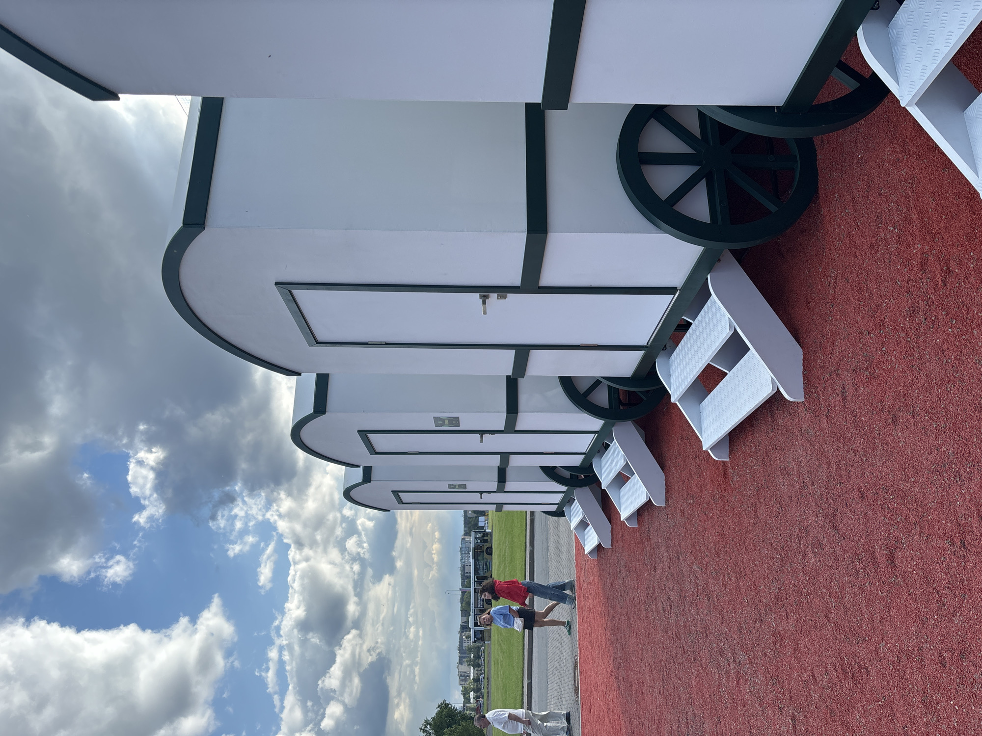 The image shows a row of white portable cabins with dark green trim and arched roofs, resembling miniature circus wagons. Each cabin has a single door with a silver handle and a set of white, slatted steps leading up to it. The cabins are positioned on a bright red, granular surface, possibly artificial turf or a specialized athletic track.
In the background, a paved walkway is visible, with three individuals walking away from the viewer. The first person is a man in a white polo shirt and light-colored pants. Behind him, a woman in a blue t-shirt and dark shorts walks alongside another woman in a red hoodie and blue jeans. Further in the background, a yellow bus with people inside is parked on a grassy field. In the distance, buildings and trees are visible against a sky with dramatic, partly cloudy skies, featuring both bright white cumulus clouds and darker, gray formations. The overall scene appears to be an outdoor public space, possibly a park or event venue.
While the image itself does not provide direct clues to its location in Saint Petersburg, Russia, such temporary structures can be found in various public spaces and parks throughout the city, particularly during events or festivals.