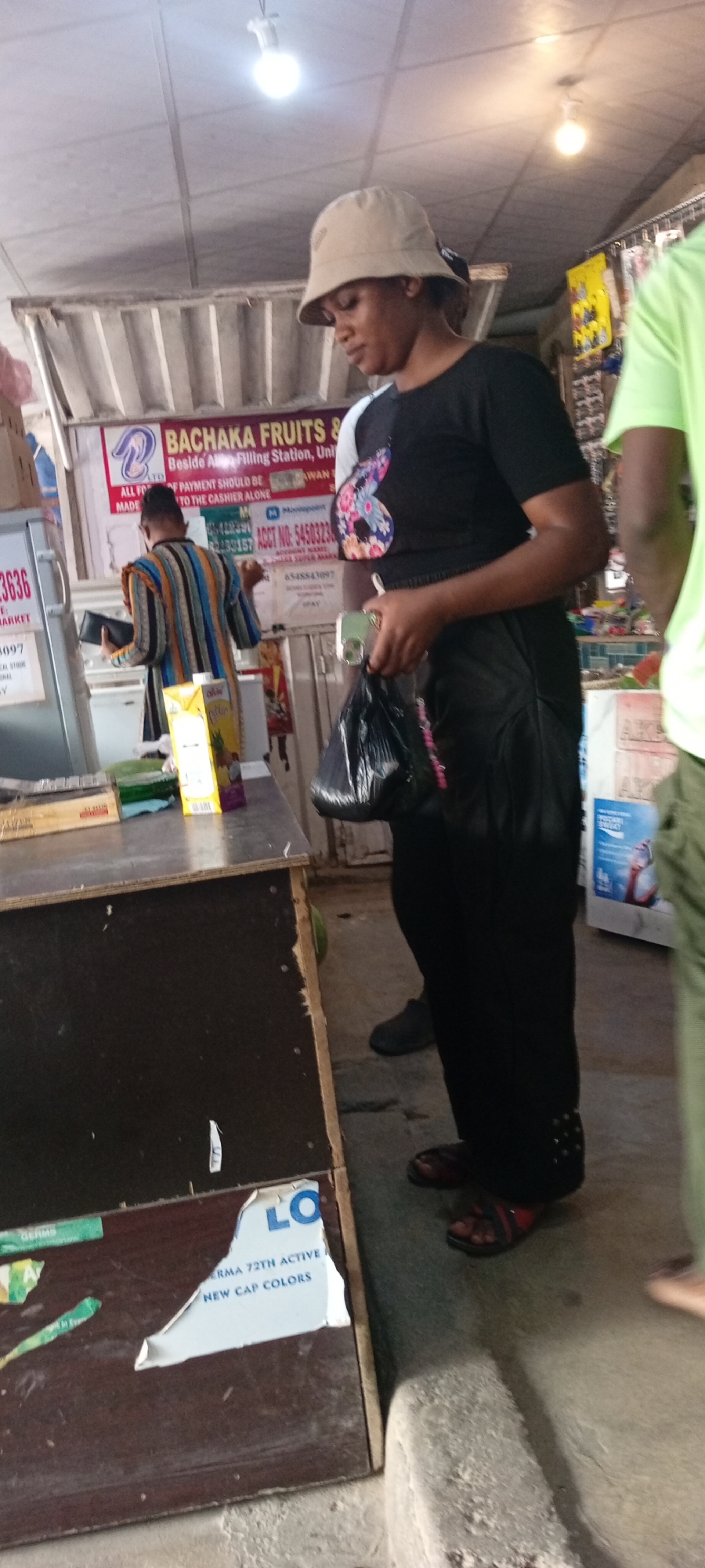 In Aviation Quarters, Nigeria, a woman wearing a black t-shirt with a colorful floral design and a light-colored bucket hat is standing near a stall. She is holding a black plastic bag in her right hand and a cell phone in her left. The stall appears to be a food or general goods vendor, with a sign above that reads "BACHAKA FRUITS &". A carton of juice is visible on the counter. Another person, partially visible in a green shirt, stands to the right. The scene is lit by overhead fluorescent lights and natural light filtering in from the open front. The ground is concrete, and there are various posters and items on display in the background.