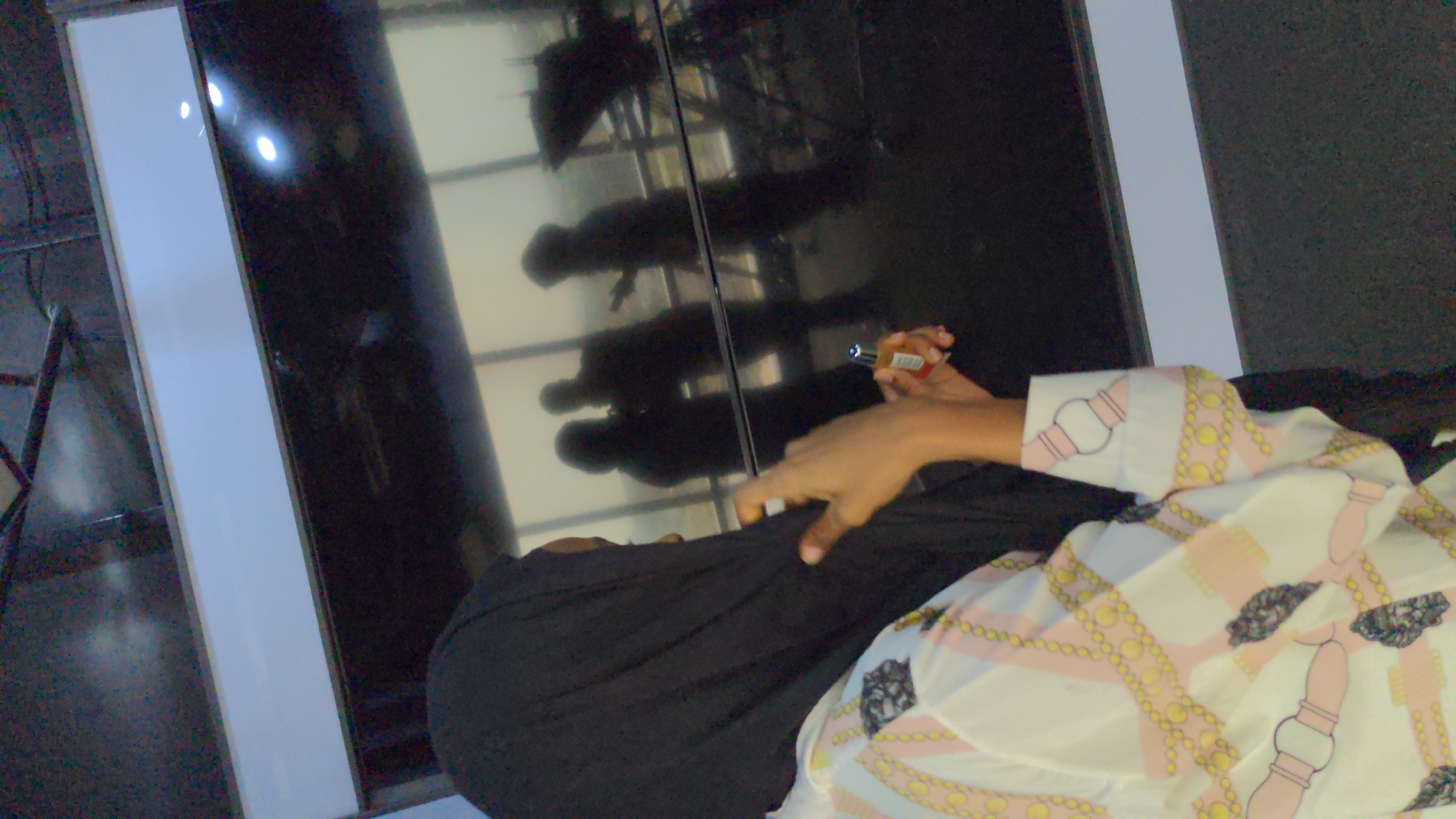 This image captures an indoor scene, likely within a modern public building in Abuja, Nigeria. The foreground is dominated by a partially visible person and a large, dark, reflective surface that fills much of the background.
In the right foreground, a person is seen wearing a white, long-sleeved shirt adorned with a repeating pattern of goldish-yellow chains and pink belt-like motifs. A dark, possibly black, garment covers their lower body or lap. The person's right hand is prominent, holding a small, red and silver electronic device that appears to be a vape pen, indicated by a glowing light at its tip, suggesting it's in use.
The background is dominated by a vast, dark, and highly reflective panel, which could be a polished floor, a wall, or a large screen. This surface acts as a mirror, showing clear, dark silhouettes of several individuals standing or moving, possibly above or in front of the reflective plane. At least three distinct human forms are discernible in these reflections. Bright, circular lights from the environment are also reflected on this dark surface. A striking vertical white strip runs along the edge of the reflective panel, contributing to a modern, structured aesthetic. In the far left of the frame, a dark, angled metal structure is visible, possibly part of a railing or furniture. The image is taken from a slightly tilted perspective. No explicit text is visible.