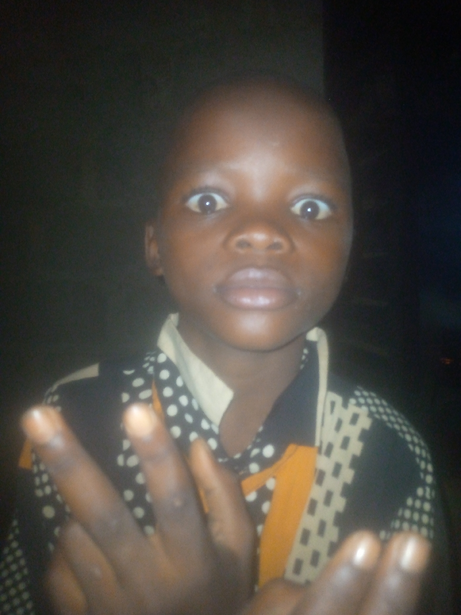 A close-up image shows a child with dark skin, large, wide eyes, and a surprised expression. The background is dark and indistinct. The child is wearing a shirt with a black and white patterned design and a yellow accent. The child's hand is held up, fingers pointing towards the viewer. There is no information in the picture to verify the location in the city of Ikorodu, Nigeria.
