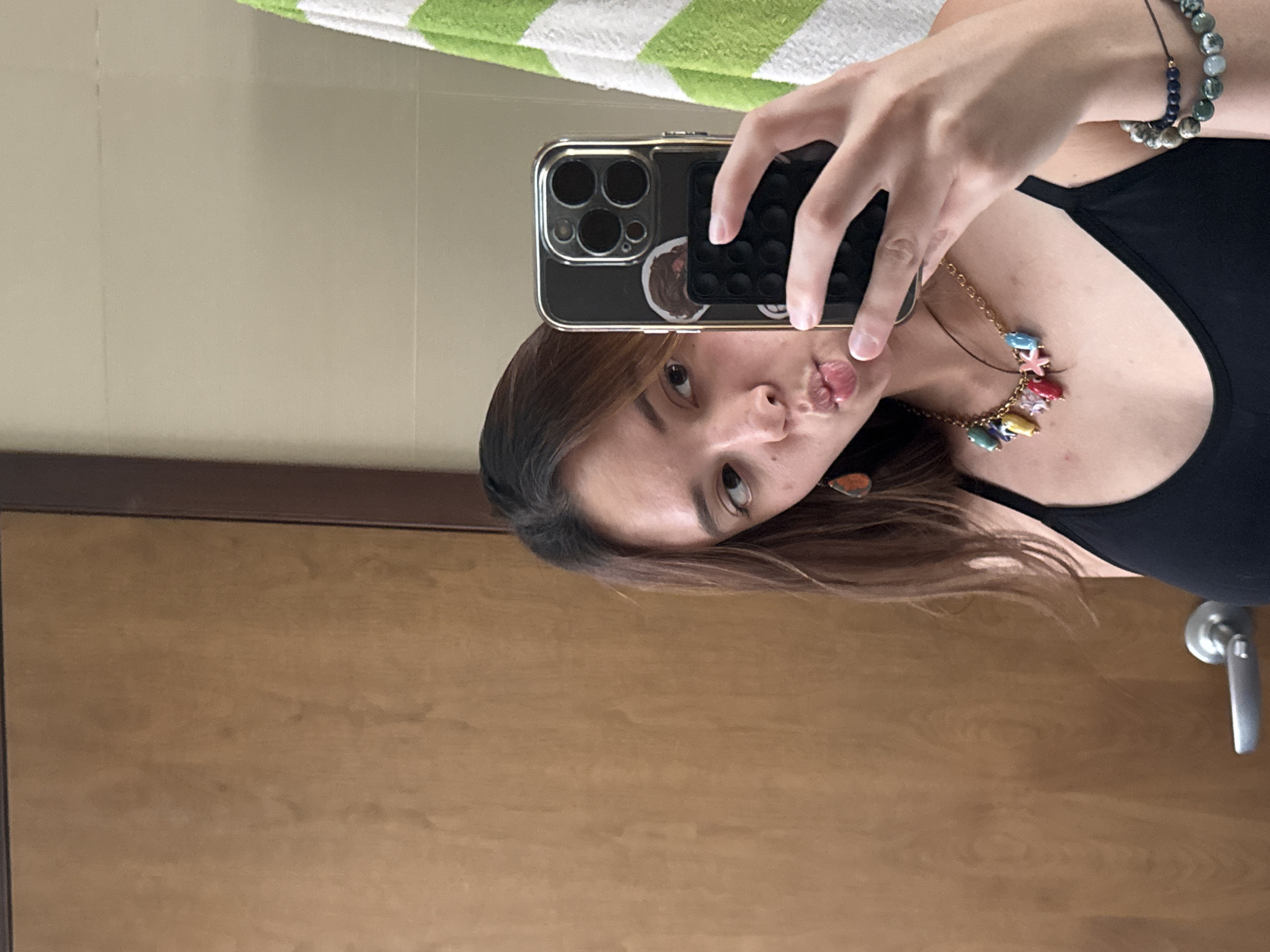 A person with medium-length brown hair is taking a selfie with an iPhone that has a black case with a gold-colored trim. The phone obscures most of their face, but their right eye, nose, and lips are visible. They are wearing a black tank top and a necklace with colorful charms. A bracelet with blue and gray beads is on their right wrist. The background includes a door with a silver handle, a wall with light beige tiles, and a green and white striped towel or cloth. The image appears to be taken indoors, possibly in a bathroom. No specific details indicate the location is San Bernardino Tlaxcalancingo, Mexico.