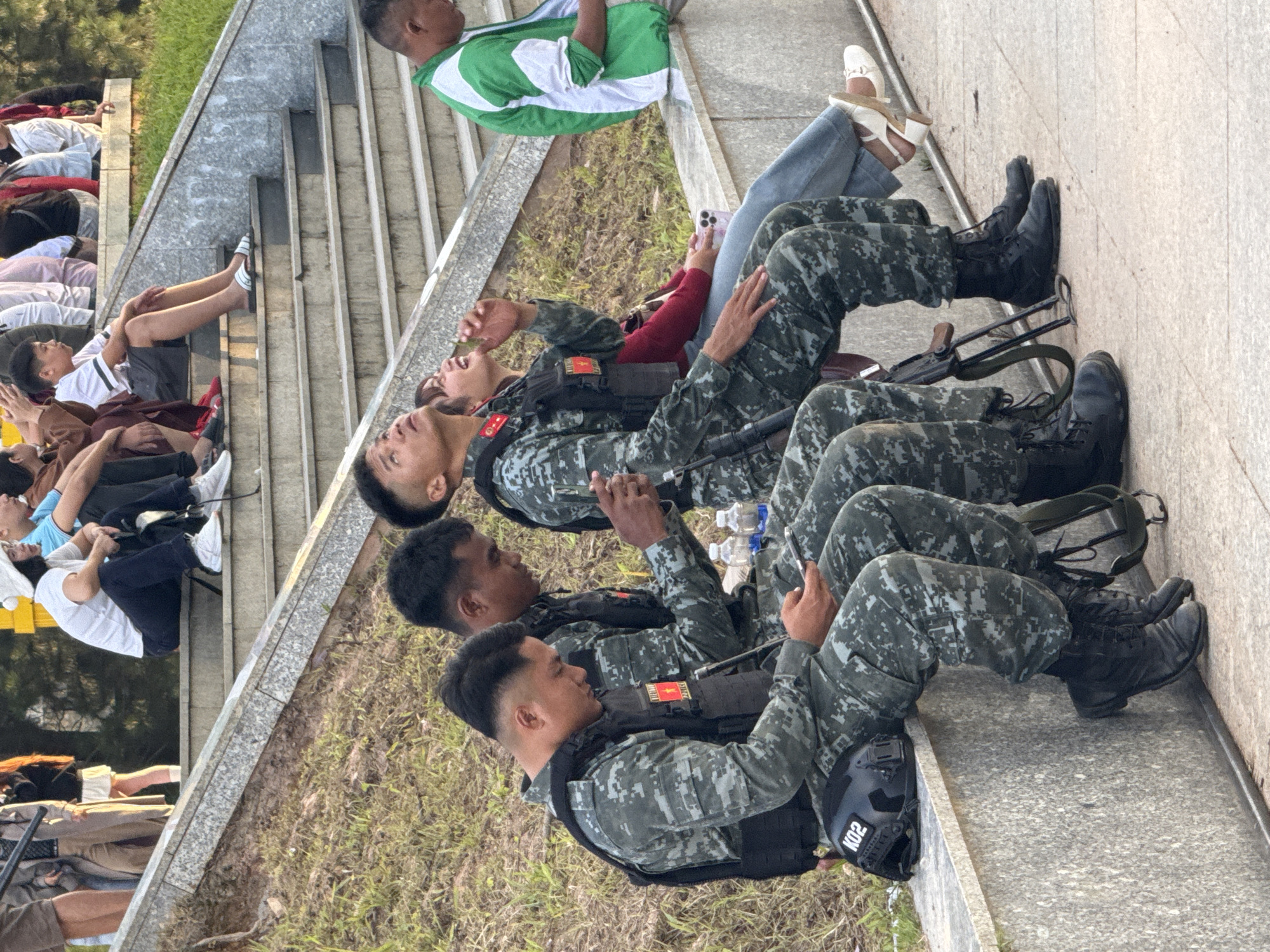 Four individuals, three in military-style camouflage uniforms and tactical vests, are seated on a concrete ledge outdoors. The individuals in uniform are focused on their mobile phones, while the fourth person, a woman in civilian clothing, is looking upwards and smiling. Two rifles are propped between two of the uniformed individuals. Behind them, a set of concrete stairs is visible, with other people seated on them. The scene appears to be at the Da Lat Flower Park in Da Lat, Vietnam, given the characteristic tiered seating and greenery often found in such public spaces in the city. The lighting suggests it is daytime.