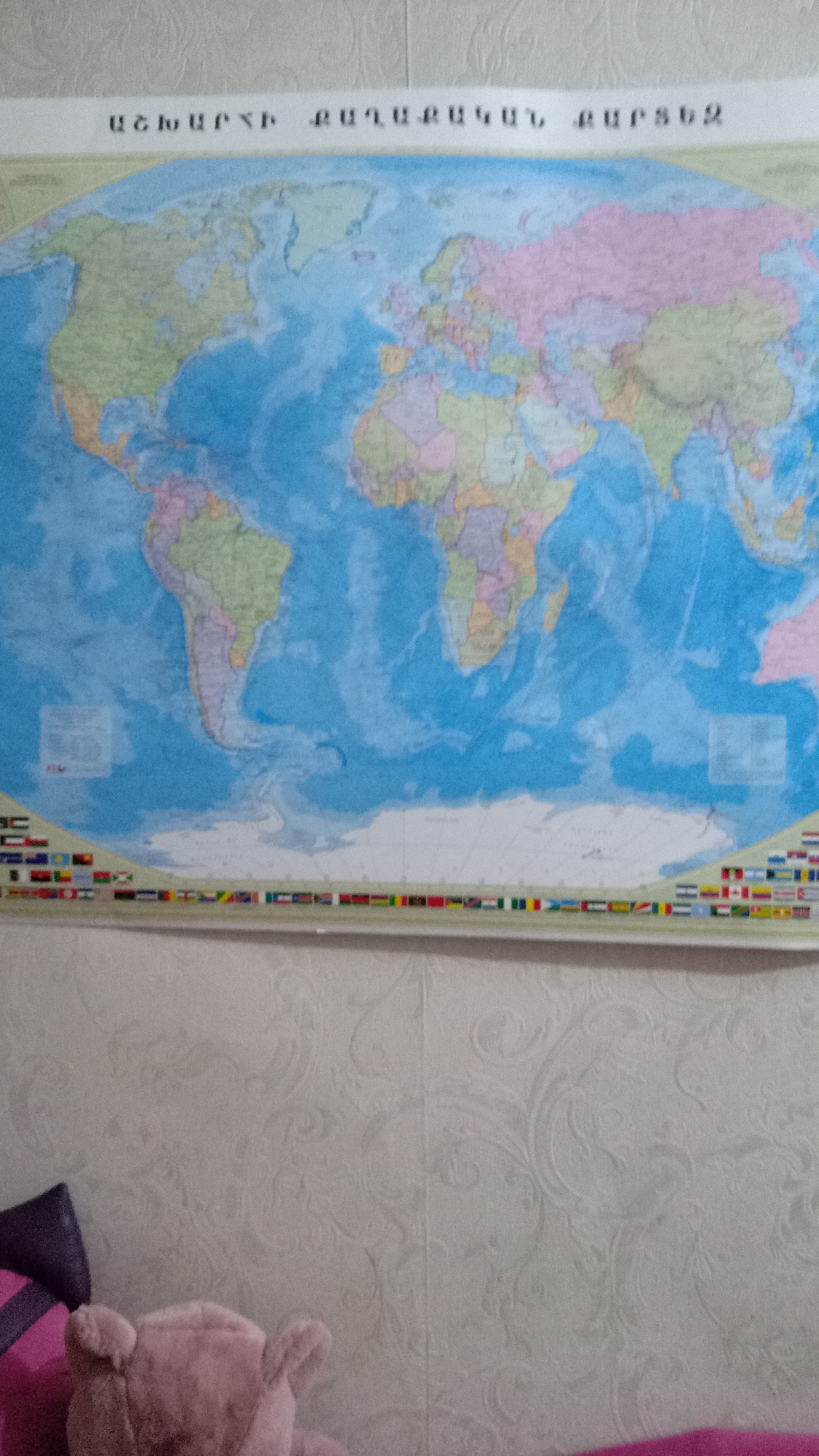 The image shows a world map hanging on a wall with patterned wallpaper. The title of the map, written in Armenian, appears to be "WORLD POLITICAL MAP". Below the map is a row of flags representing various countries. In the foreground, partially visible, are a teddy bear and a pillow. The location in the city of Yerevan, Armenia, cannot be determined from the image.