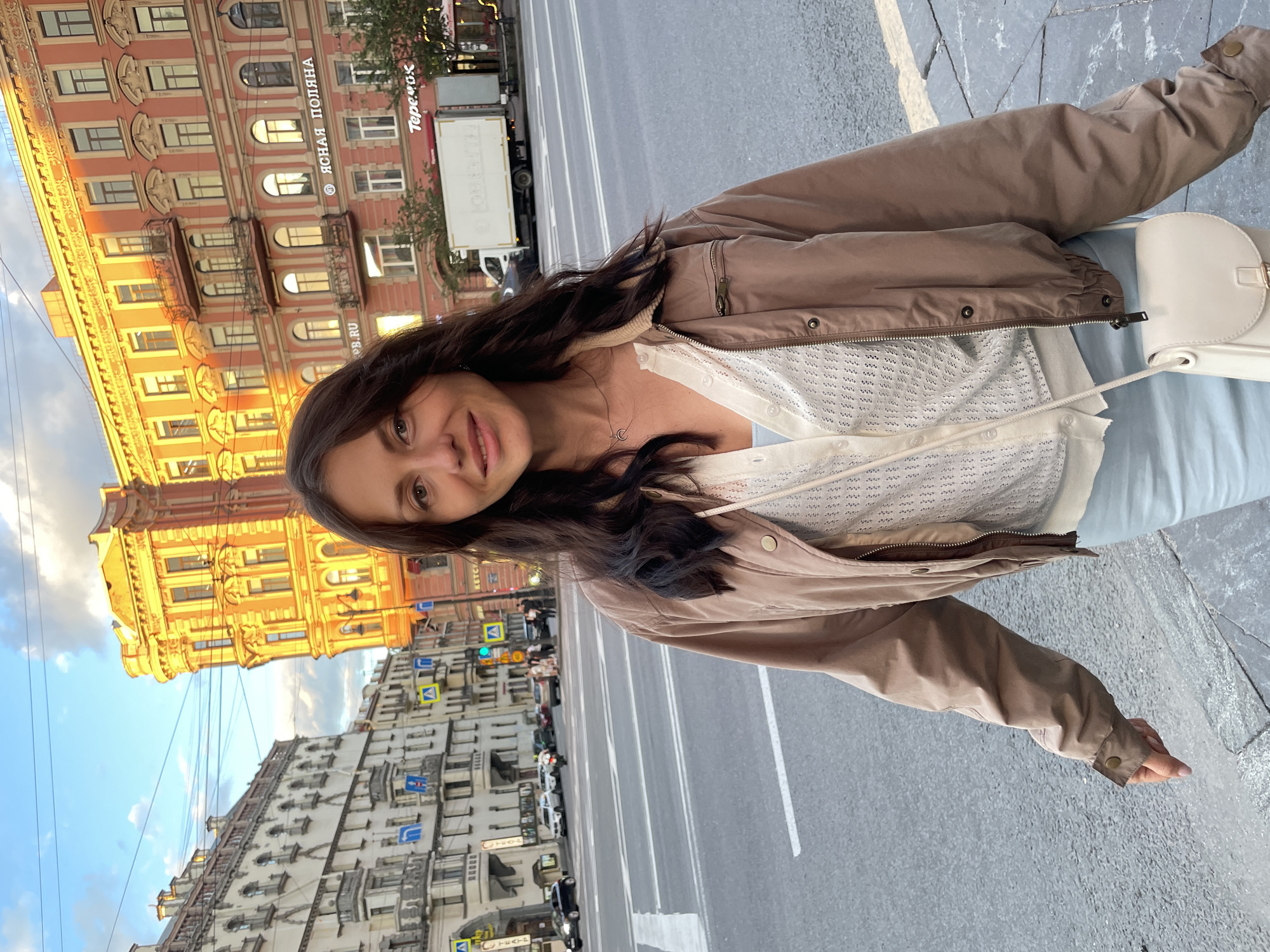 A woman with long, dark hair smiles at the camera in a medium shot. She is wearing a light blue top, a cream-colored cardigan, and a brown bomber jacket. She has a small white crossbody bag with a gold clasp. The woman is standing on a sidewalk next to a street with a crosswalk. In the background, there are buildings with orange and beige facades, some of which have architectural details like balconies and arched windows. A truck and several cars are visible on the street. The sky is a mix of blue and clouds. The scene appears to be in Saint Petersburg, Russia, likely near Nevsky Prospekt, given the architectural style and the presence of street signs.