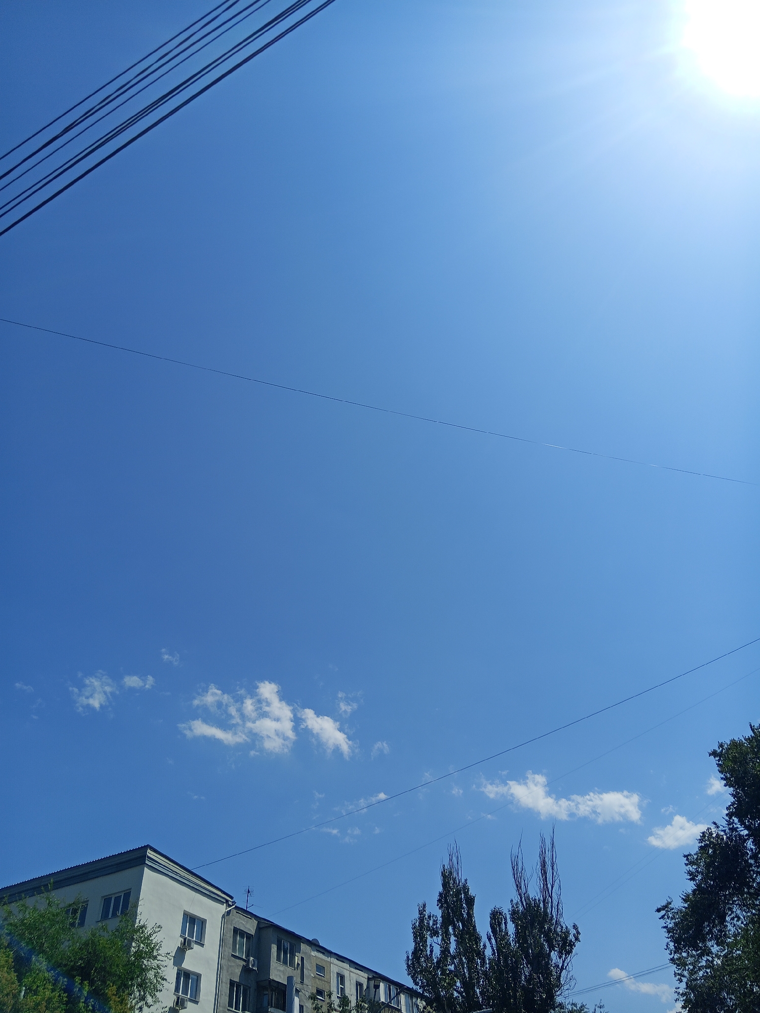 The image depicts a clear blue sky on a sunny day. The sun is visible in the upper right quadrant, casting bright rays. Several power lines stretch across the sky, some of which are quite prominent in the upper left. In the lower portion of the image, the rooftops of apartment buildings are visible, along with some trees. The buildings appear to be multi-story residential structures with windows and some visible air conditioning units. The overall scene suggests an urban environment on a bright, clear day, likely in a residential district.