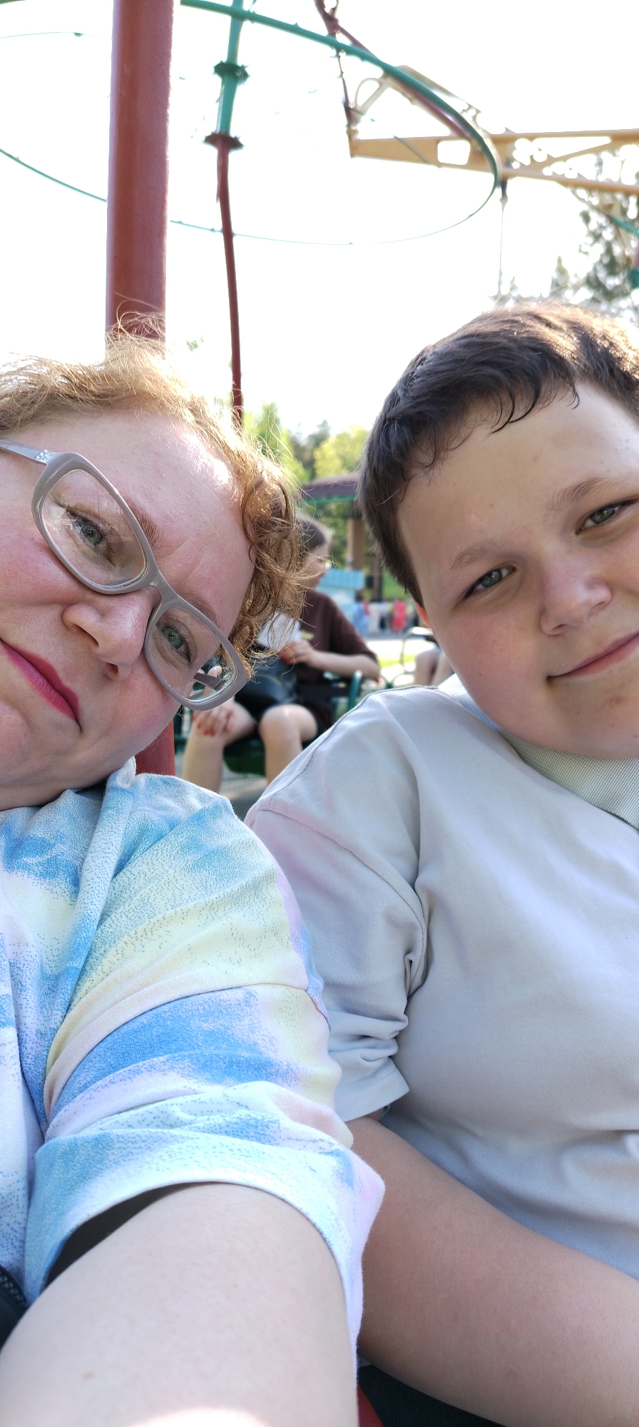 A selfie shows a woman and a boy sitting together in an outdoor setting. The woman, on the left, has red hair and wears glasses and red lipstick. She is wearing a blue and white tie-dye shirt. The boy, on the right, has dark hair and is wearing a white shirt. Behind them, indistinct figures sit on benches in what appears to be a park. A tall, red pole structure is visible in the background. The photograph was taken in the city of Chaikovsky, Russia.