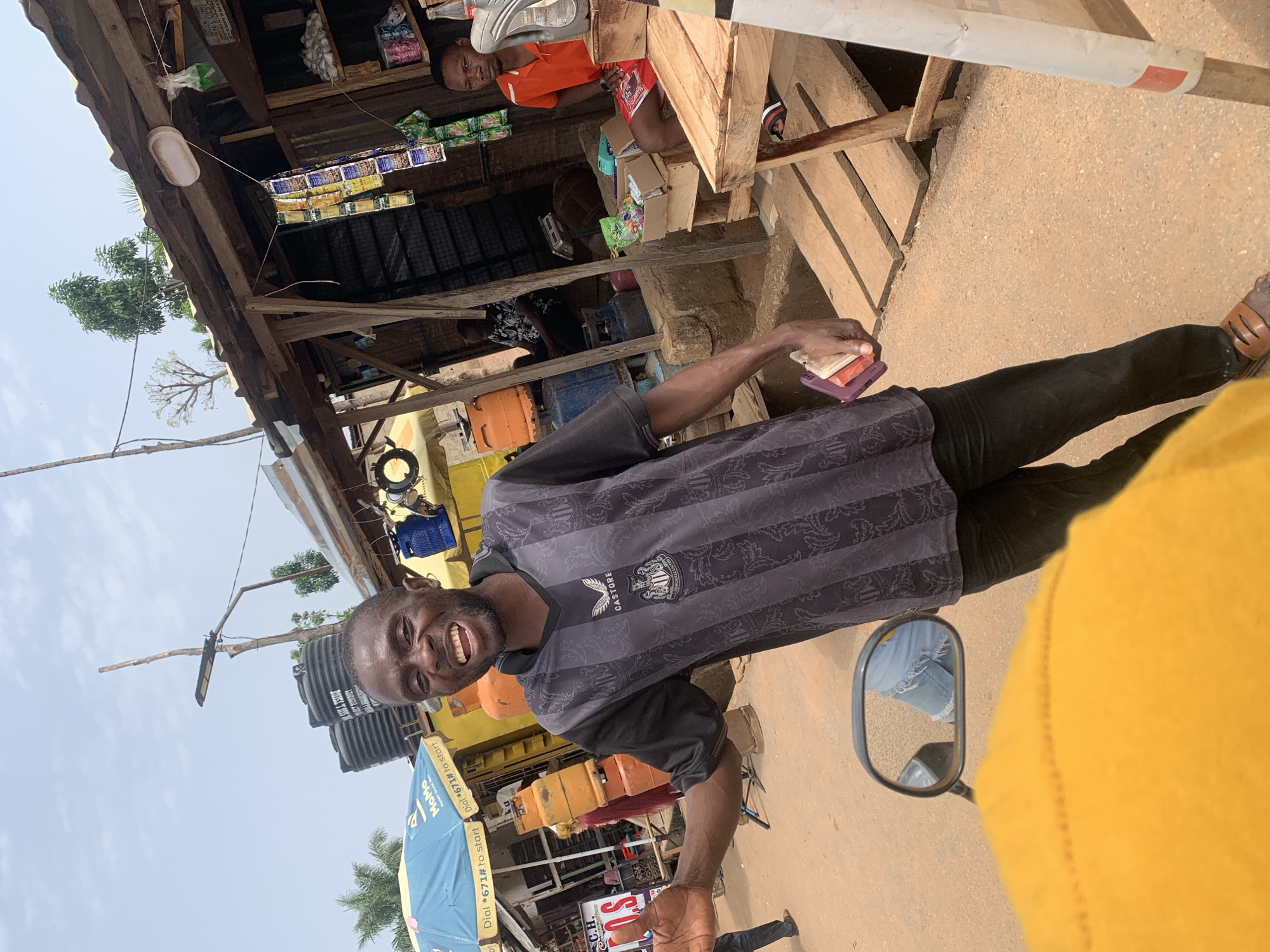 The image captures a scene in Akwanga, Nigeria. A smiling man wearing a black and grey Newcastle United soccer jersey stands in the street. He's holding a purple phone case and several small, possibly paper, items in his hand.
Behind him, a small roadside shop is visible, displaying various items for sale. A person in an orange shirt sits inside the shop. Two water tanks are visible above the shop's awning, along with a street light. A motorcycle side mirror is visible, reflecting a person's leg wearing ripped jeans. A yellow shirt is in the foreground.
The environment suggests a typical street scene in a Nigerian town, with a combination of commercial activity and everyday life taking place.