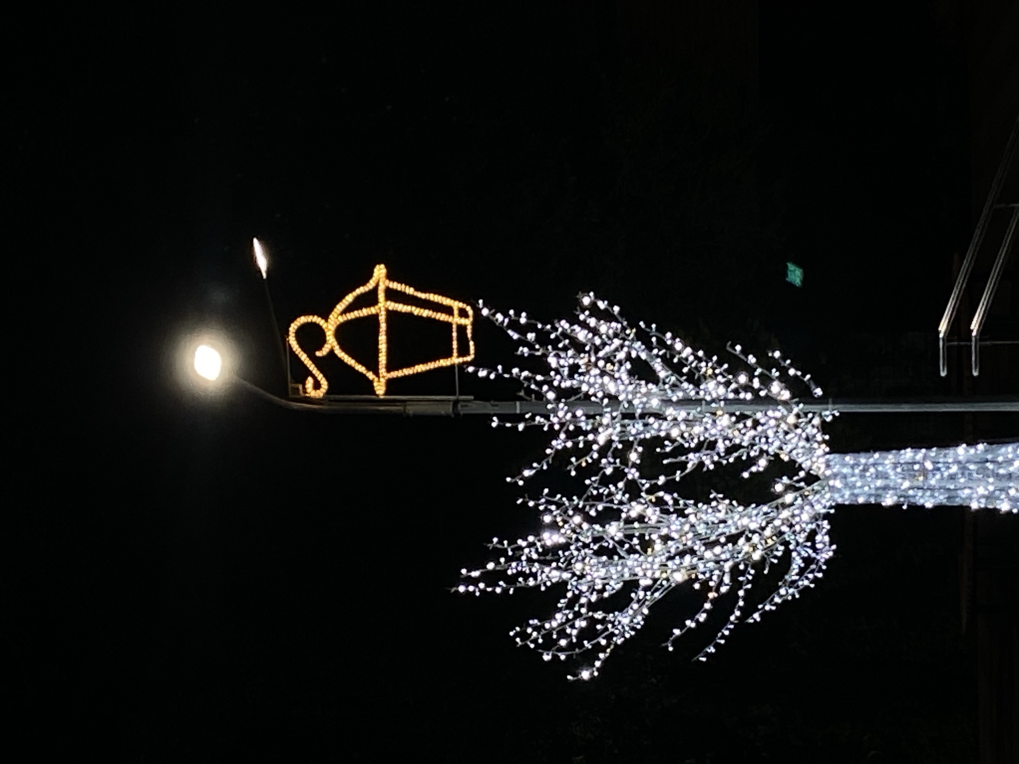 This is a nighttime photograph taken in Tolyatti, Russia, focusing on outdoor decorative lighting. A large, artificial tree covered in white LED lights stands prominently on the left side of the frame. To its right, a lamppost is topped with a festive, lantern-shaped light decoration outlined in yellow LEDs. A bright, white streetlamp is also visible on the lamppost, casting light downwards. The background is dark, indicating nighttime. In the lower right corner, a metal railing for stairs can be seen. A small, green, square object is suspended in the air in the middle-right portion of the image, its purpose or nature unclear.