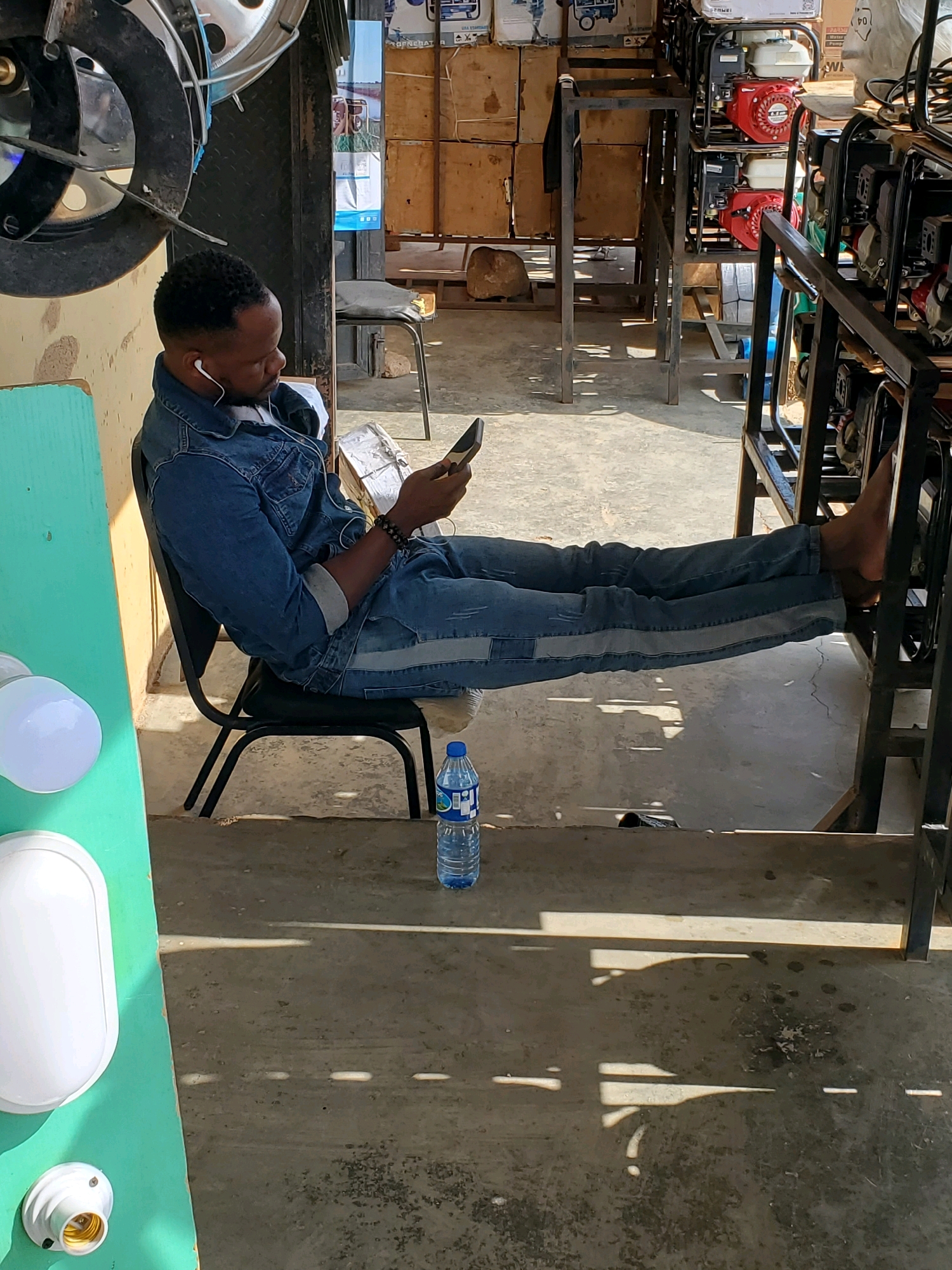 A Black man wearing a denim jacket and jeans sits on a black chair, his legs extended, with his feet resting on a metal structure. He is holding a smartphone in his right hand and appears to be looking at it. White earbuds are in his ears. Next to him on the floor is a bottle of water. The scene appears to be an outdoor or semi-outdoor area with a concrete floor. In the background, there are stacks of wooden crates and several portable generators. Sunlight casts shadows on the ground. The presence of generators and crates suggests a commercial or storage area, possibly a shop or warehouse. The environment is informal.
