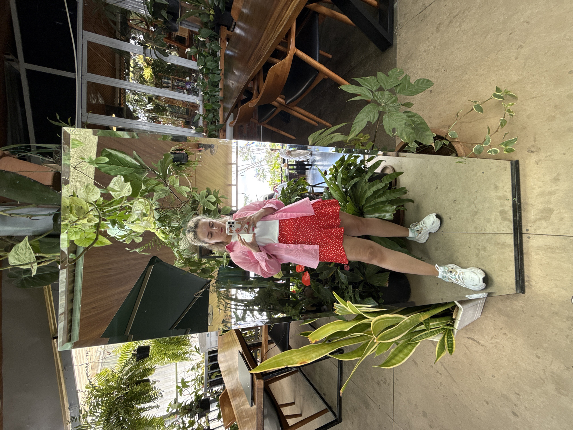 A medium, full shot captures a woman reflected in a large mirror in what appears to be a plant-filled interior space. The scene is likely located in Da Lat, Vietnam, based on the plant-filled interior space, common in the city. The woman is holding a phone in her left hand, appearing to take a photo of herself in the mirror. The woman is wearing a white top, a red dress with white polka dots, and a light pink button-down shirt. She is wearing white sneakers. The mirror shows reflections of plants, furniture, and architectural details of the interior. The floor is a neutral tile. To the right of the mirror are potted plants, and below the mirror is a sansevieria plant in a gray pot. Through the mirror's reflection, more furniture and greenery are visible. A large window structure is seen behind the furniture. The lighting in the scene is natural and diffused, illuminating the subject and surroundings evenly. The image seems casual, suggesting a spontaneous moment captured by the subject.