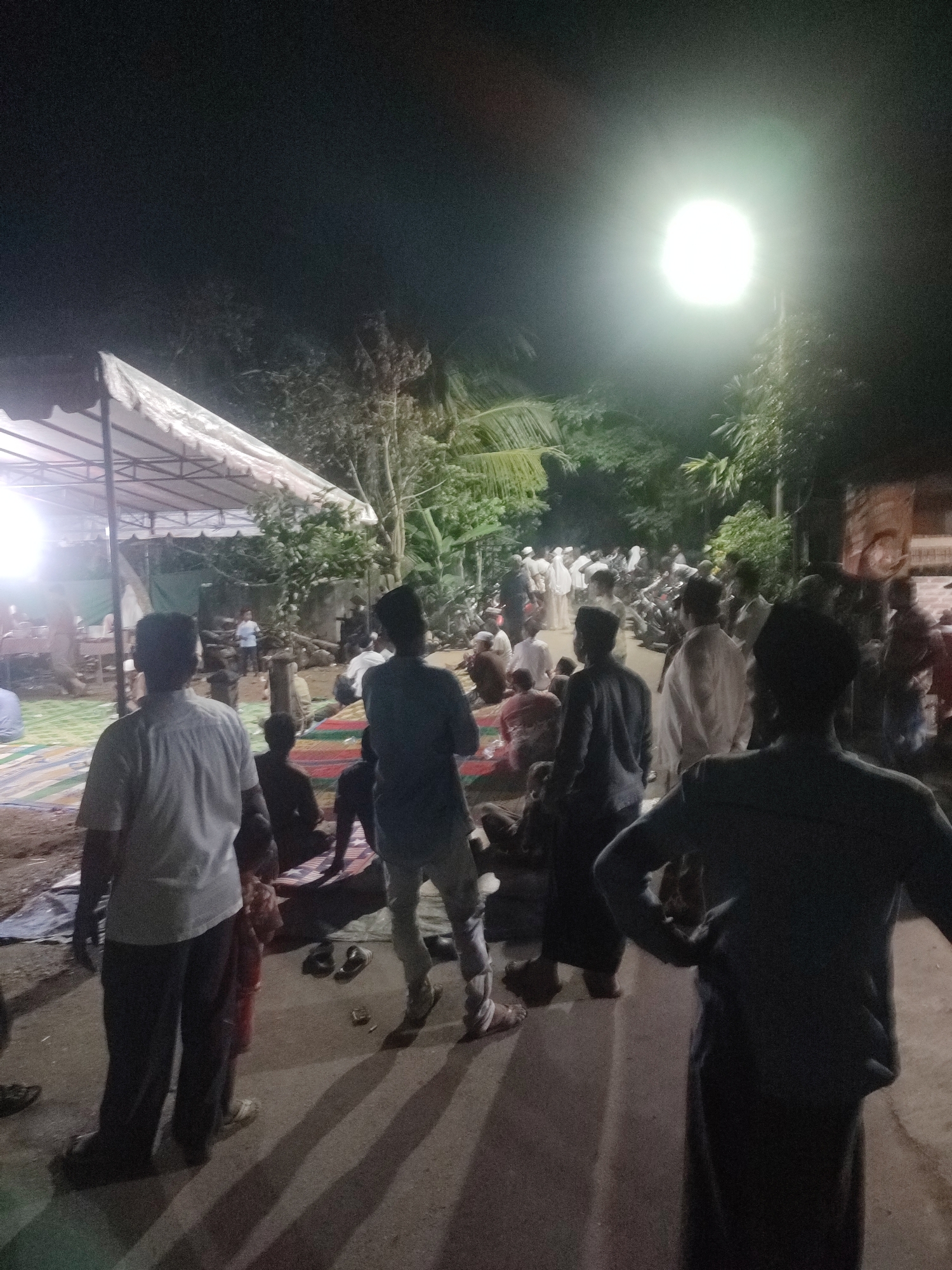 This media file depicts a gathering of people at night, possibly for an event or religious service, in Langkuta, Indonesia. The scene is illuminated by a bright overhead light source, creating a stark contrast with the surrounding darkness and casting long shadows. A white tent structure is visible on the left, suggesting a temporary shelter or venue for the occasion. Many individuals are dressed in traditional attire, with several men wearing skullcaps. Some people are seated on mats laid out on the ground, while others are standing and interacting. The atmosphere appears to be one of community and shared activity, though specific emotions are difficult to ascertain due to the low lighting and distance. The backdrop includes foliage and trees, indicating an outdoor setting, possibly in a rural or semi-rural area. The ground appears to be unpaved, with some dirt and sand visible. The presence of multiple people suggests a significant gathering. No visible text is discernible in the image.