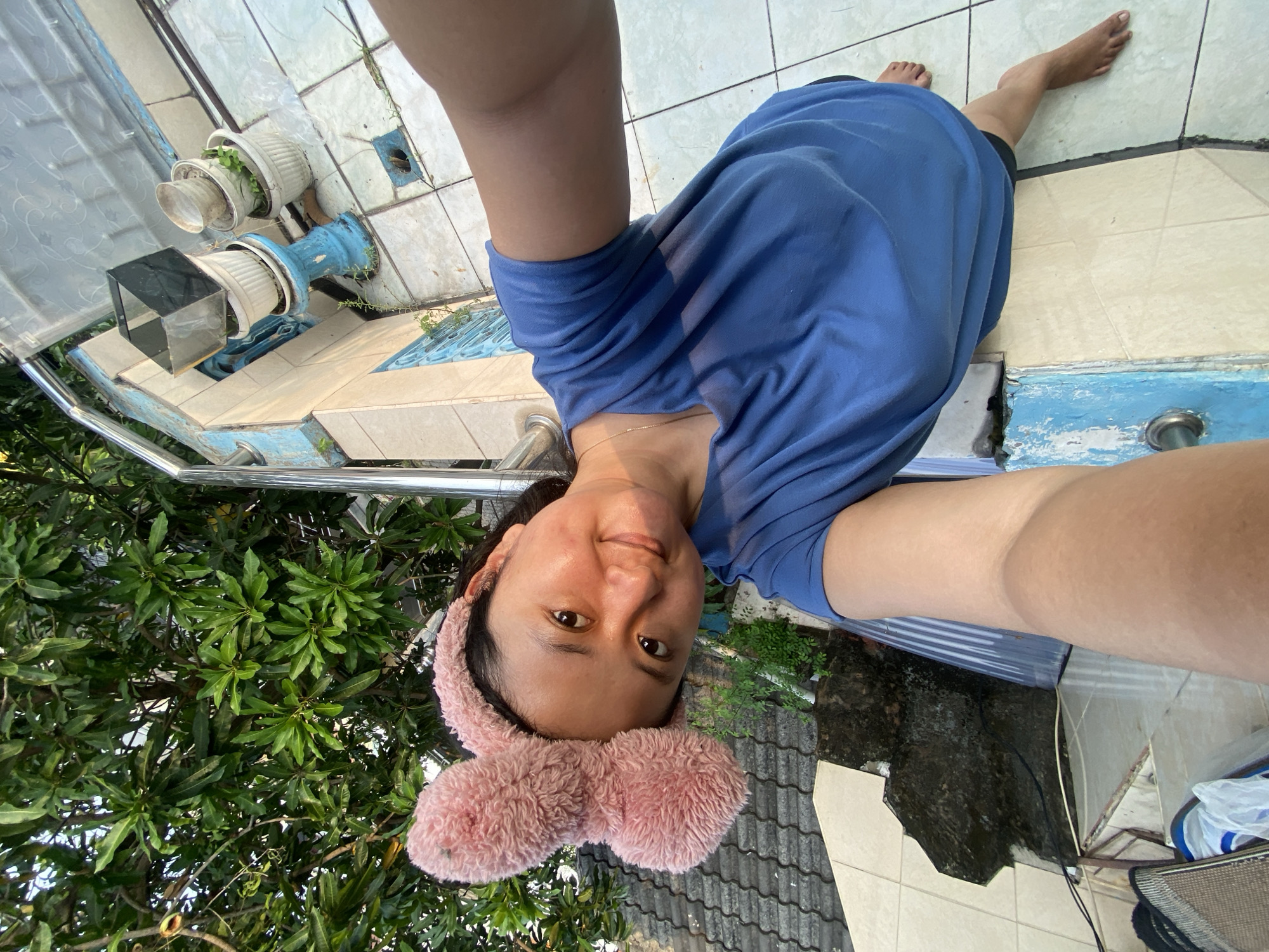 The image is a high-angle selfie taken by a young adult woman in an outdoor setting, likely a patio or backyard in Gelam, Indonesia. The woman is seen from the chest up, smiling subtly at the camera. She has dark hair and wears a light blue t-shirt and a fluffy pink headband resembling bunny ears. Her bare feet are visible towards the upper right, resting on the tiled ground.
The ground is covered with light-colored, possibly beige or off-white, ceramic tiles, some with blue decorative patterns. The tiles show signs of age, with visible cracks and small weeds growing through them. A low, light blue painted structure, possibly a wall or planter, runs along one side of the tiled area. Several white plastic pots containing green plants are placed on this structure. One pot has additional objects, including a cylindrical item and a square, dark-colored object on top.
Dense green foliage, characteristic of tropical trees or bushes, forms a lush background, suggesting a natural outdoor environment. A dark gray or black tiled roof and a wet, dark patch of ground are also visible in the background. Metal railings and a translucent corrugated sheet, possibly part of a canopy or roof, are present in the scene.
The bright, natural light indicates daytime, likely morning or afternoon, given the soft shadows. The overall atmosphere is relaxed, with the woman appearing calm and content. No visible text is present in the image.