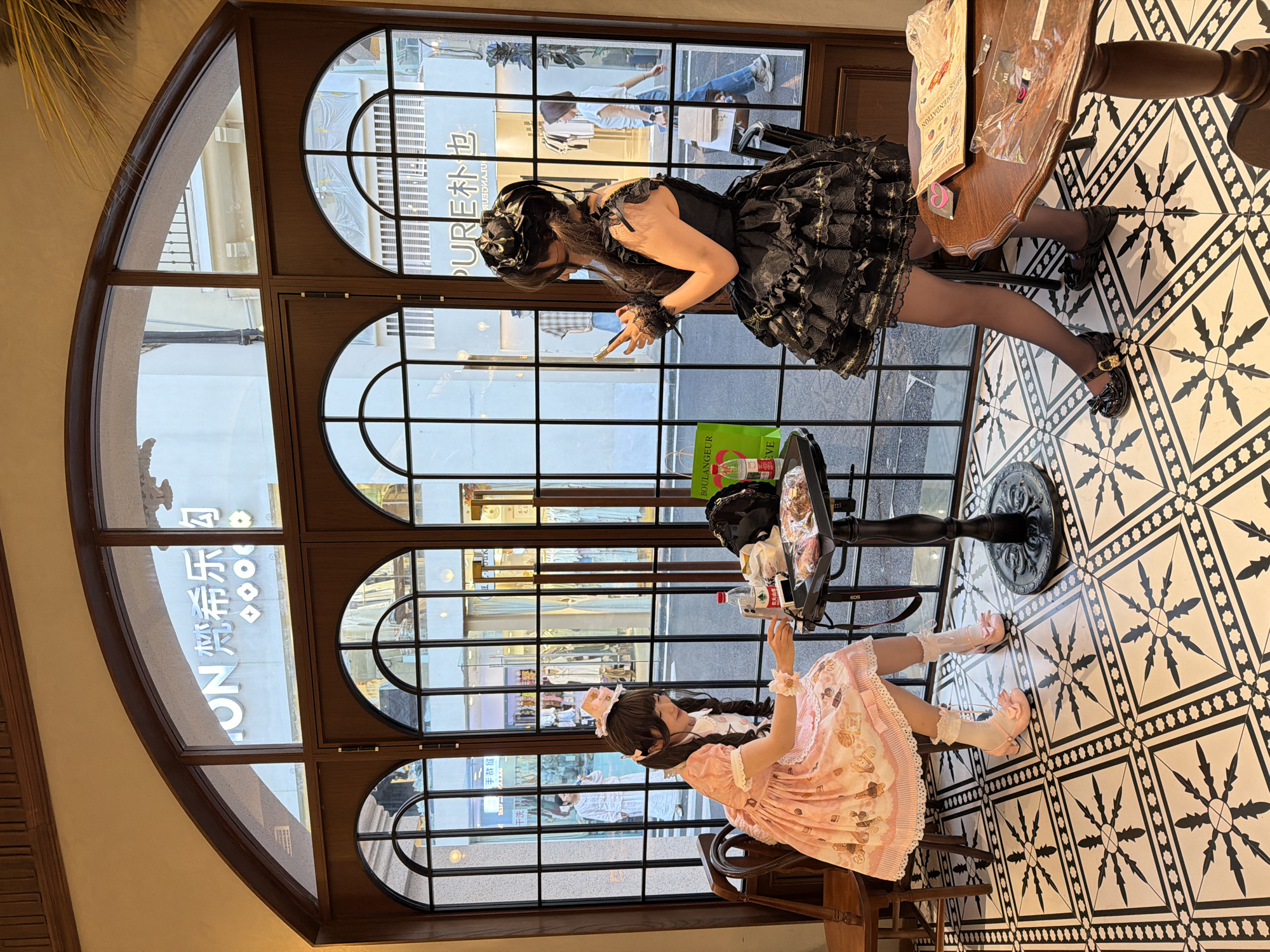 Here is a detailed description of the image:
**Scene:** The photograph captures an interior scene with two women dressed in Lolita fashion, presumably in a restaurant or cafe. The location appears to be in the Chongqing High-tech Zone, China, based on the Chinese characters visible on a window storefront.
**People:**
* Two women are prominently featured in the image.
* One woman is wearing a black Lolita dress with lace and ornate detailing, black tights, and black shoes. She is standing by a small table and counter.
* The second woman is wearing a pink Lolita dress with frills and lace, light socks with lace detailing, and pink shoes. She is seated on a chair and appears to be interacting with something on the table, like a drink.
* There are blurred figures visible outside the window, suggesting other people walking on the street.
**Objects:**
* The interior is decorated with distinctive black and white geometric patterned tiles.
* There are tables and chairs scattered around.
* A small table has items on it, including drinks.
* Outside the windows, storefronts are visible. The words "PURE" in English and Chinese characters are visible.
**Actions and Interactions:**
* The women seem to be engaged in some activity, possibly taking photos.
* The scene suggests a social gathering or outing.
**Environment:**
* The interior appears to be a restaurant or cafe.
* Large arched windows provide natural light and a view of the street.
* The architectural design and decor suggest a modern setting.