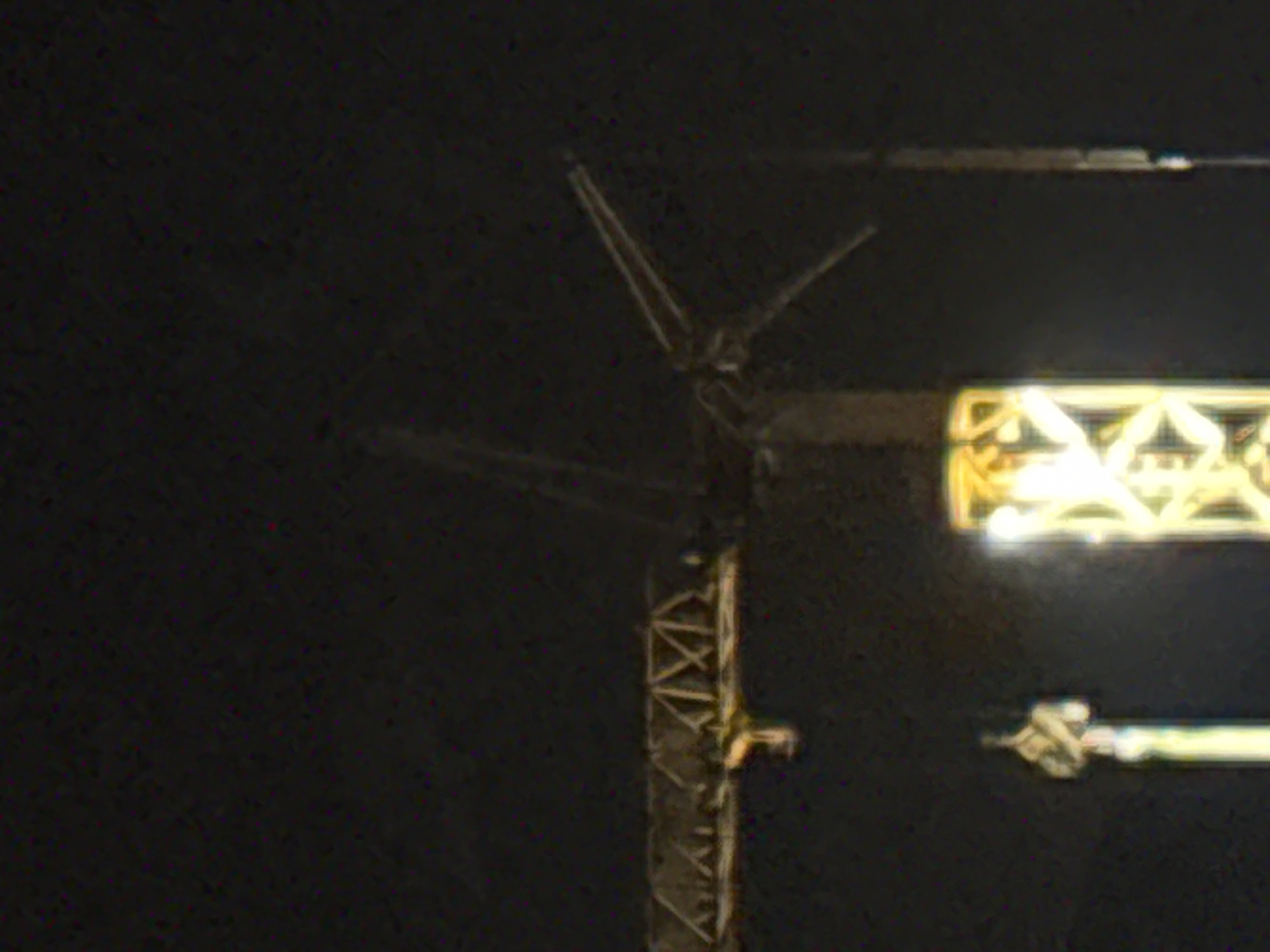 The image depicts a construction crane at night in Boreti, Montenegro. The crane is a prominent object in the foreground, its lattice structure illuminated by artificial lights. The arm of the crane extends towards the right, supporting a hook and cable. In the background, a brightly lit structure, possibly another part of the construction or a building, is visible. The scene is dark, suggesting it is nighttime. There is no visible text in the image. The overall impression is one of ongoing construction work in a coastal area, given the mention of Boreti, Montenegro, which is a coastal town.