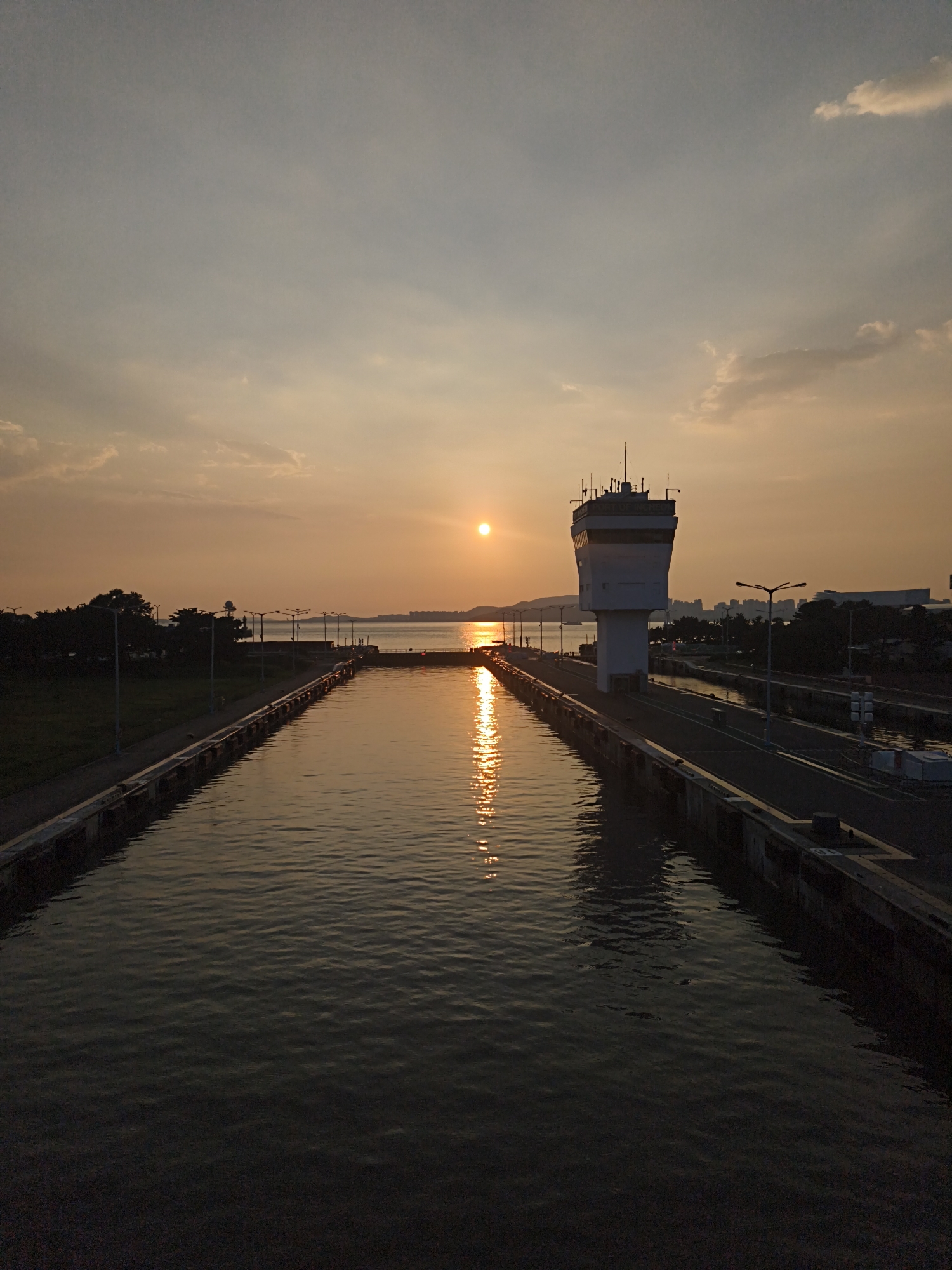 This is a vertical, eye-level, full shot of a sunset over a lock in Incheon, South Korea. The sun is setting on the right side of the frame, casting a warm orange glow across the sky and reflecting on the calm water of the lock. The lock is a rectangular channel with concrete walls on either side, leading into the distance. A tall, white control tower stands on the right side of the lock, with antennas on top. Lights are visible along the sides of the lock, and trees and buildings can be seen in the distance on the left and right horizons. The water is dark and still, with the exception of the shimmering reflection of the sun. There are no people visible in the image. The scene is tranquil and serene, capturing the beauty of nature at dusk. The location is confirmed as the Incheon Lock, a vital infrastructure for maritime traffic in the city.