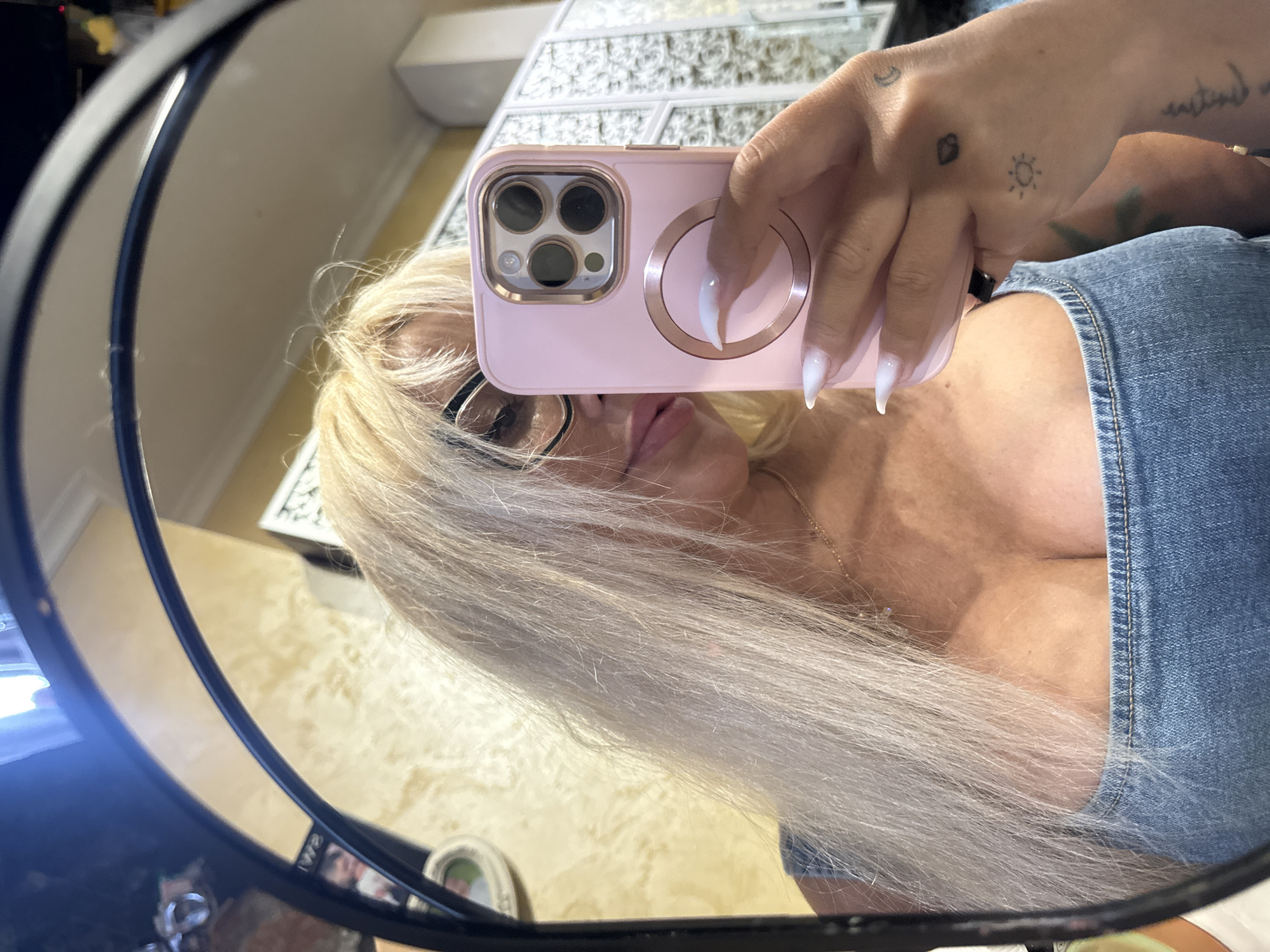 A woman with long blonde hair, wearing glasses and a denim top, takes a mirror selfie in what appears to be a residential setting. She is holding a pink phone with a gold accent around the camera lens. Her nails are long and white. Tattoos are visible on her hand, including a sun and a moon. The background shows textured walls and decorative screens. The image does not provide specific information to identify the location as Odesa, Ukraine.