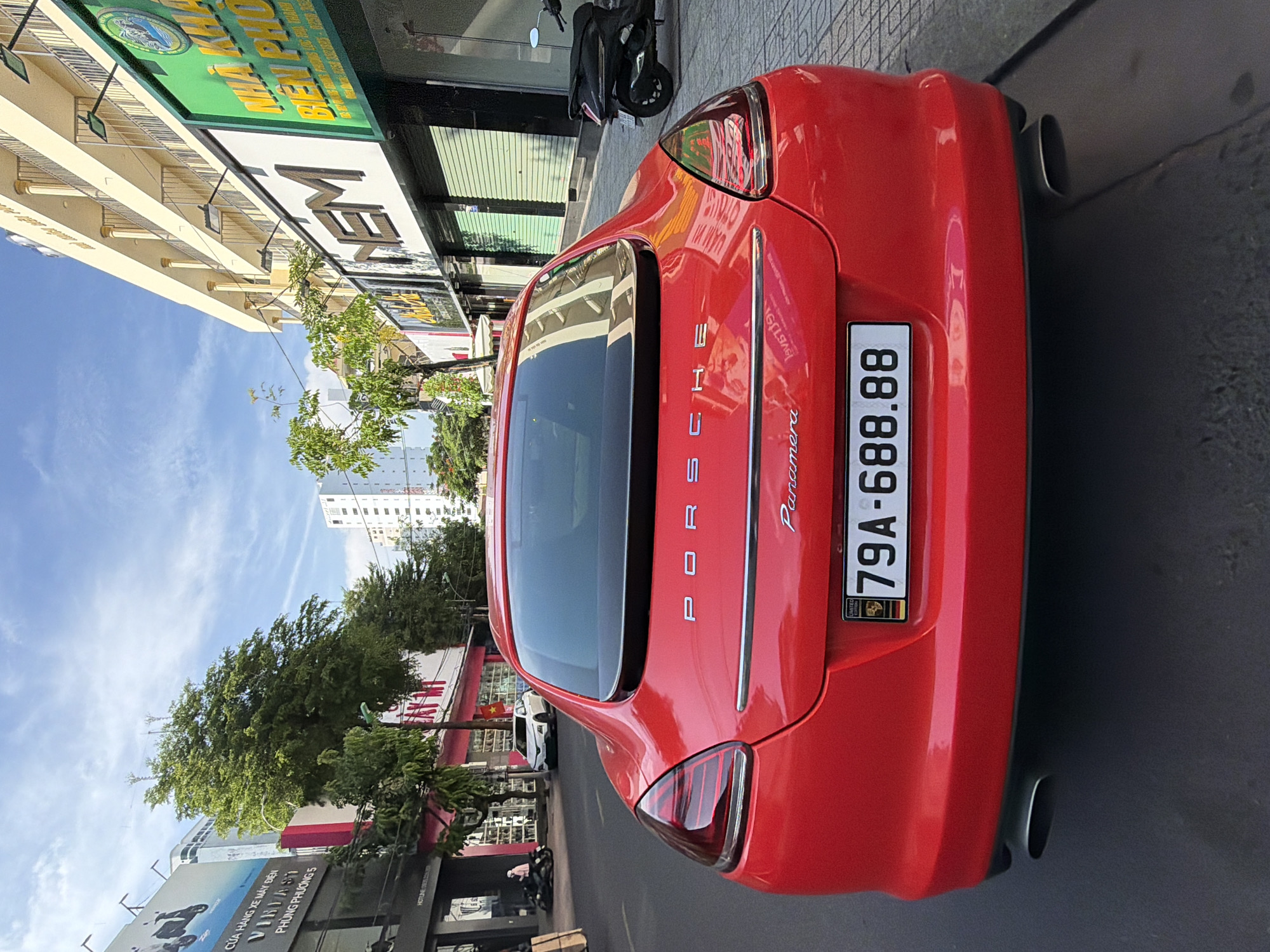 A red Porsche Panamera is parked on a city street in Nha Trang, Vietnam. The car is facing away from the camera, and its rear is prominently displayed. The license plate reads "79A-688.88". The car is parked on the right side of the street, with a sidewalk next to it. The sidewalk is paved with grey tiles. On the sidewalk, there are several buildings with shops on the ground floor. The buildings are modern and colorful, with signs in Vietnamese. There are trees planted along the sidewalk. In the background, there are more buildings, including a tall white building. The sky is blue with some white clouds. A black scooter is parked on the sidewalk to the right of the car. A person is seen walking on the sidewalk to the left of the car. The scene appears to be daytime, with good lighting.