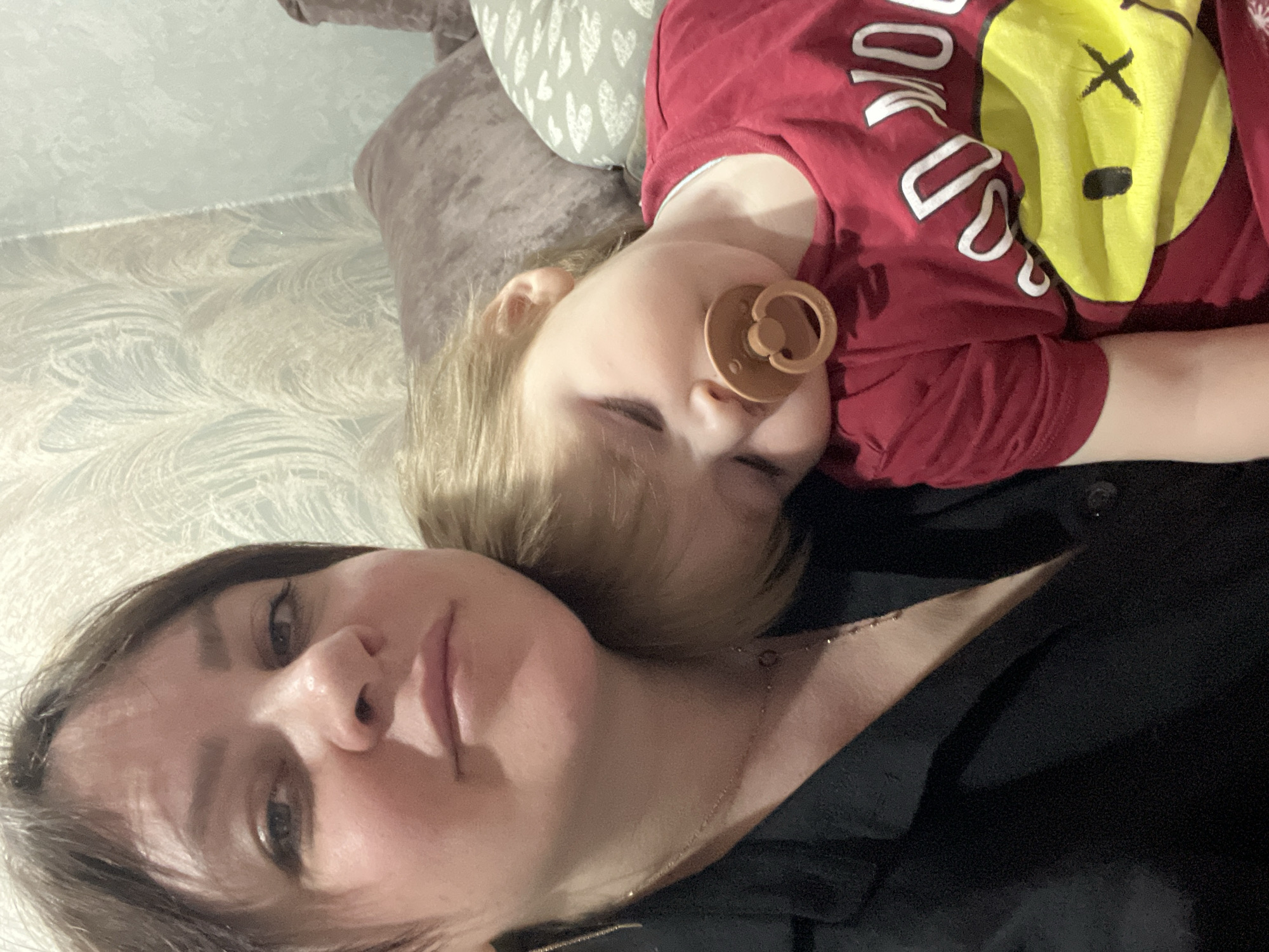 A photograph depicts a woman and child lying on a couch. The child, wearing a red t-shirt with a yellow graphic and a pacifier, is asleep on the woman's chest. The woman is wearing a black shirt and a necklace. A pillow is behind them. The location is unknown, and no information indicates this is located in Noviy, Russia.