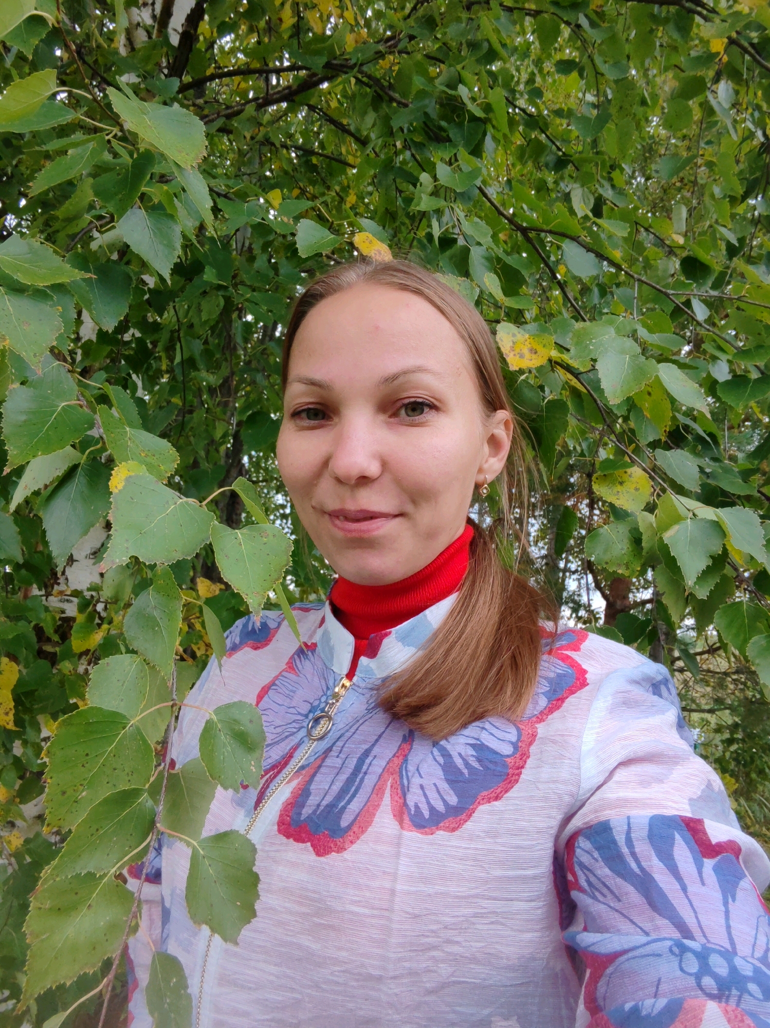 A medium shot is captured outdoors, featuring a woman with light brown hair pulled back in a ponytail and light green eyes. She is smiling and looking directly at the camera. She is wearing a red turtleneck shirt underneath a light-colored, patterned jacket with large floral designs in pink and blue. The woman is positioned in front of a backdrop of green leaves, suggesting a natural outdoor setting, possibly in a park or forest. The specific location of Чухлэм, Russia, is not visually discernible from the image itself.
