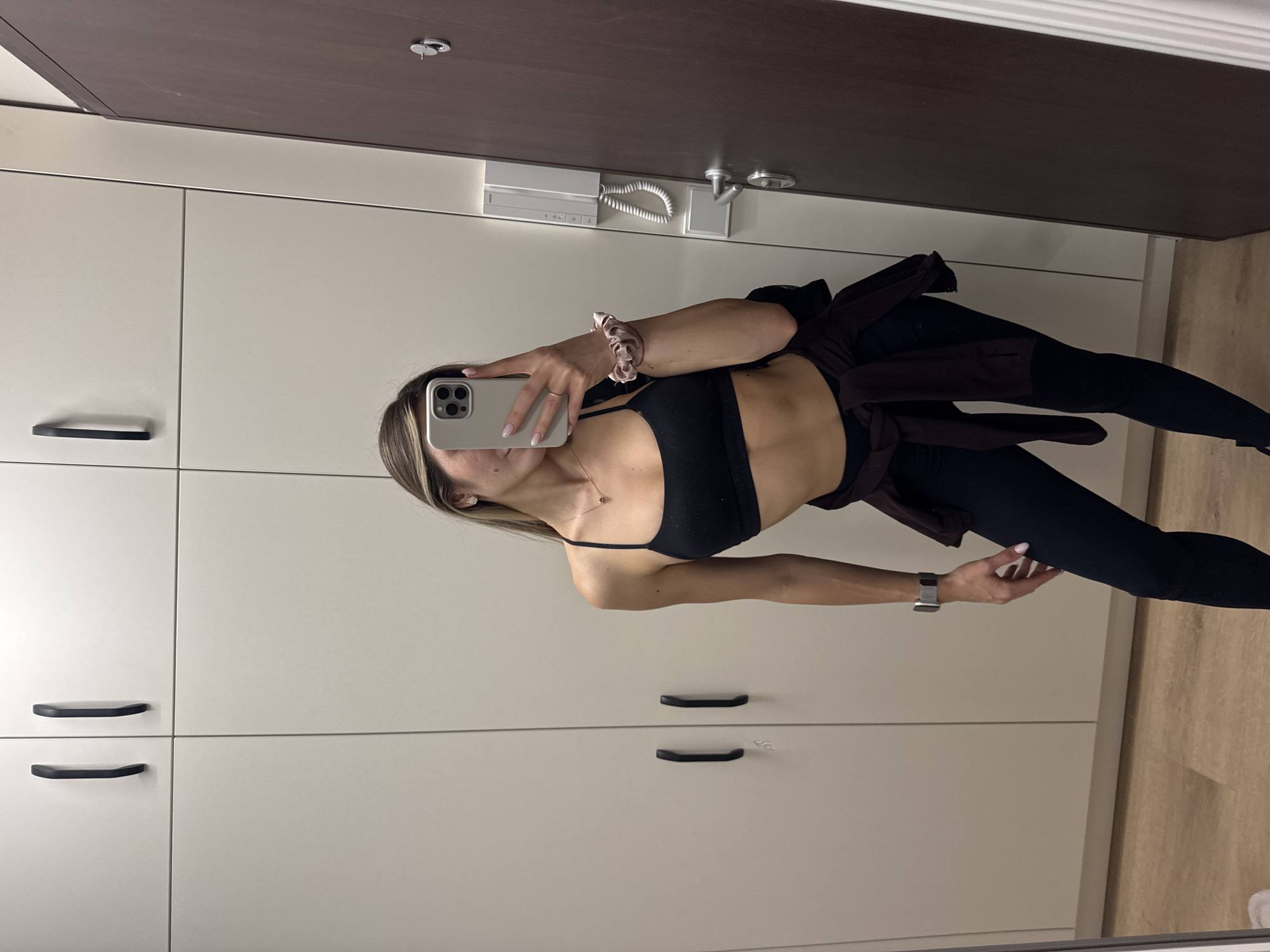 A young woman is captured in a full-body mirror selfie taken indoors, likely in Sofia, Bulgaria. She stands slightly angled, holding a light-colored smartphone with a visible camera array to take the photograph. Her long, light brown hair with blonde highlights is styled straight, falling over her shoulders.
She is dressed in activewear, consisting of a black sports bra or crop top that exposes her midriff, and black form-fitting leggings. A dark brown or burgundy jacket or sweatshirt is tied around her waist. Accessories include a light pink scrunchie on her right wrist, a thin gold-colored necklace, small stud earrings, and a silver-toned Apple Watch on her left wrist. Her fingernails are painted a light pink.
The background of the reflection reveals a modern indoor space. To her left and below, there are light-colored, possibly off-white, minimalist cabinets with sleek black horizontal handles. Above the cabinets and behind her, a dark brown wooden door with a silver handle is visible, along with a silver circular door stop or hook on the ceiling/upper door frame. An intercom system with a white coiled cord is mounted on the wall above the cabinets and below the door frame. The floor visible in the lower right portion of the image is made of light-colored wood. No discernible text is visible within the image beyond the unreadable branding on the phone or watch.