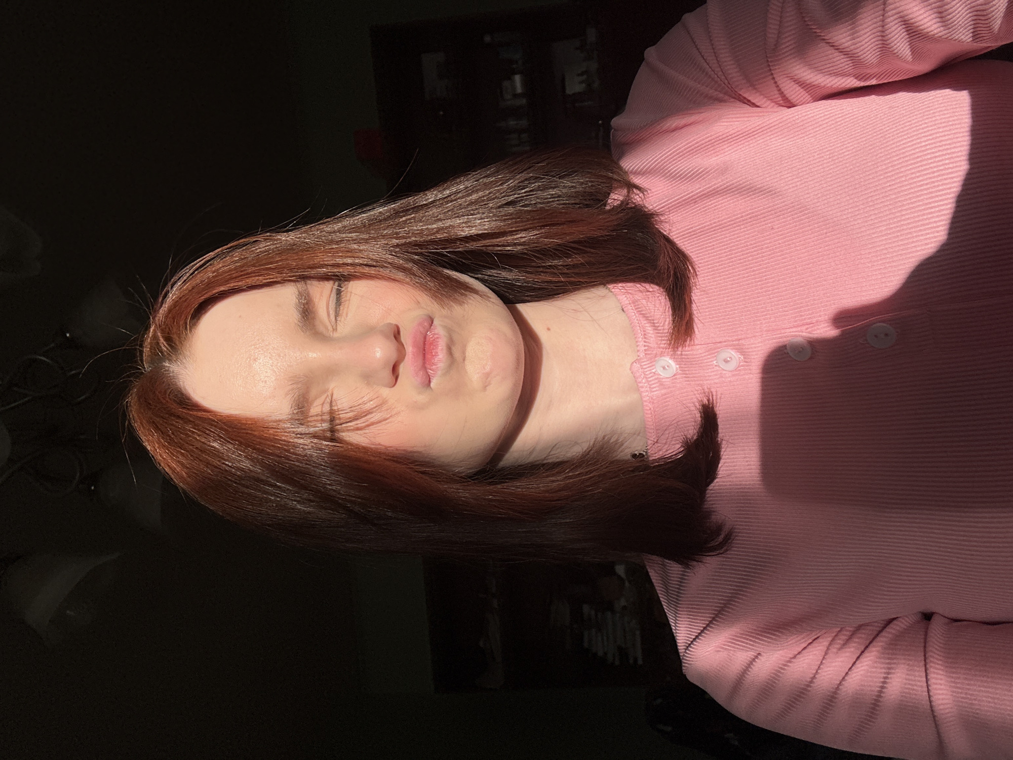 A photograph, taken in an indoor setting with a city of Unknown, Russia as a location, showcases an individual with auburn hair, wearing a pink button-down shirt with white buttons. The lighting appears to be a mix of natural light and shadows, with half of the face being brightly lit and the other half partially obscured by shadow. The individual has their eyes closed and their lips pursed in what appears to be a slight pout. The background is dark and indistinct, suggesting the presence of furniture or objects that are not easily discernible due to the lighting.