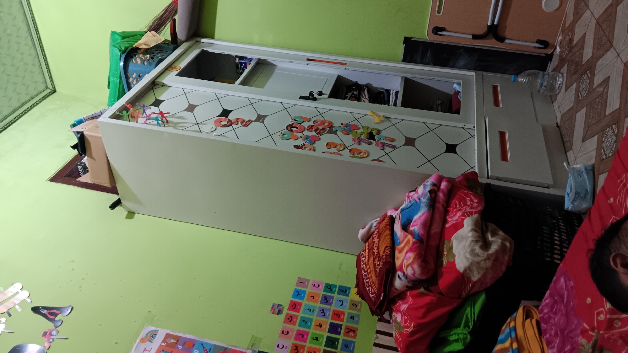 The image depicts an indoor scene, likely a bedroom or playroom, in a residential setting. The dominant colors are light green on the walls and off-white for a tall wardrobe. The wardrobe has a door with a diamond pattern and is adorned with colorful letter and number magnets. On top of the wardrobe, several items are stacked, including a cardboard box, a blue laundry basket, and green plastic bags. To the left of the wardrobe, a wall displays colorful charts with Arabic letters and some decorative cutouts of toys. In the foreground, a pile of folded blankets and textiles is stacked on a dark basket. A portion of a person, possibly a child, is visible lying down on a red patterned surface in the bottom left corner. Other objects in the scene include a plastic bottle of water on the floor and a folded brown table to the right. The flooring appears to be tiled. The overall impression is of a lived-in space, possibly a child's room, with a slightly cluttered but organized appearance. The image has a slight tilt, suggesting it was taken from an angle. The location is identified as Sonobekel, Indonesia.