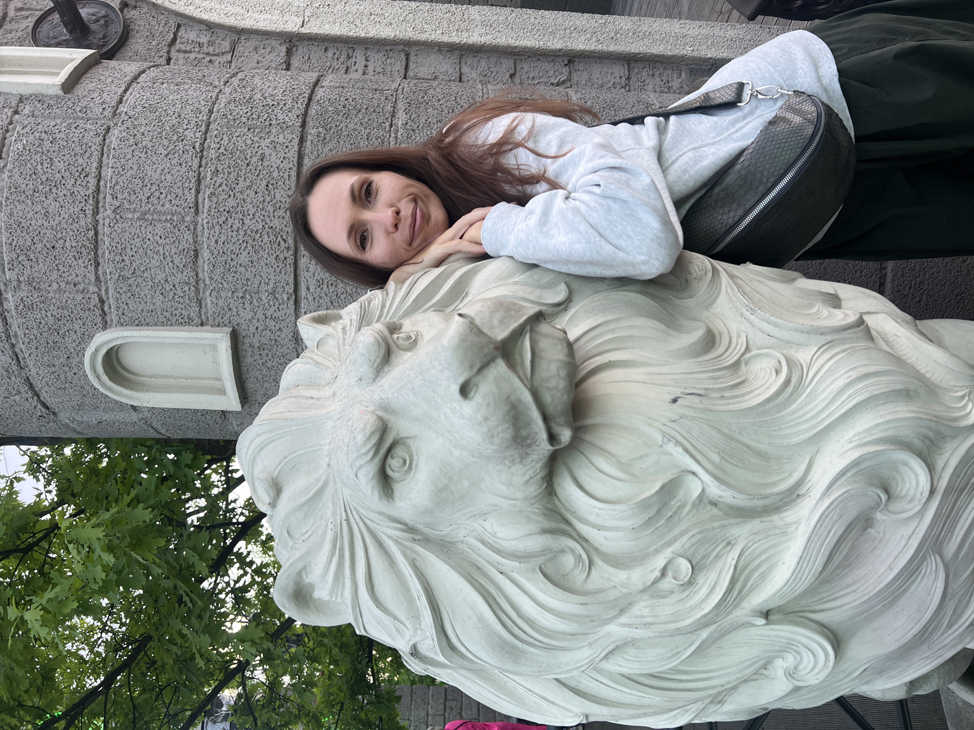 A woman is reclining on a stone lion statue in Moscow, Russia. She is fair-skinned with long brown hair and wearing a light gray sweatshirt with a dark gray snakeskin-patterned cross-body bag. She is smiling and looking at the camera with her hands resting under her chin.
The lion statue is made of white stone with intricate carvings of its mane. The statue sits against a textured gray building facade. Green trees and foliage are partially visible in the lower-left corner of the image.