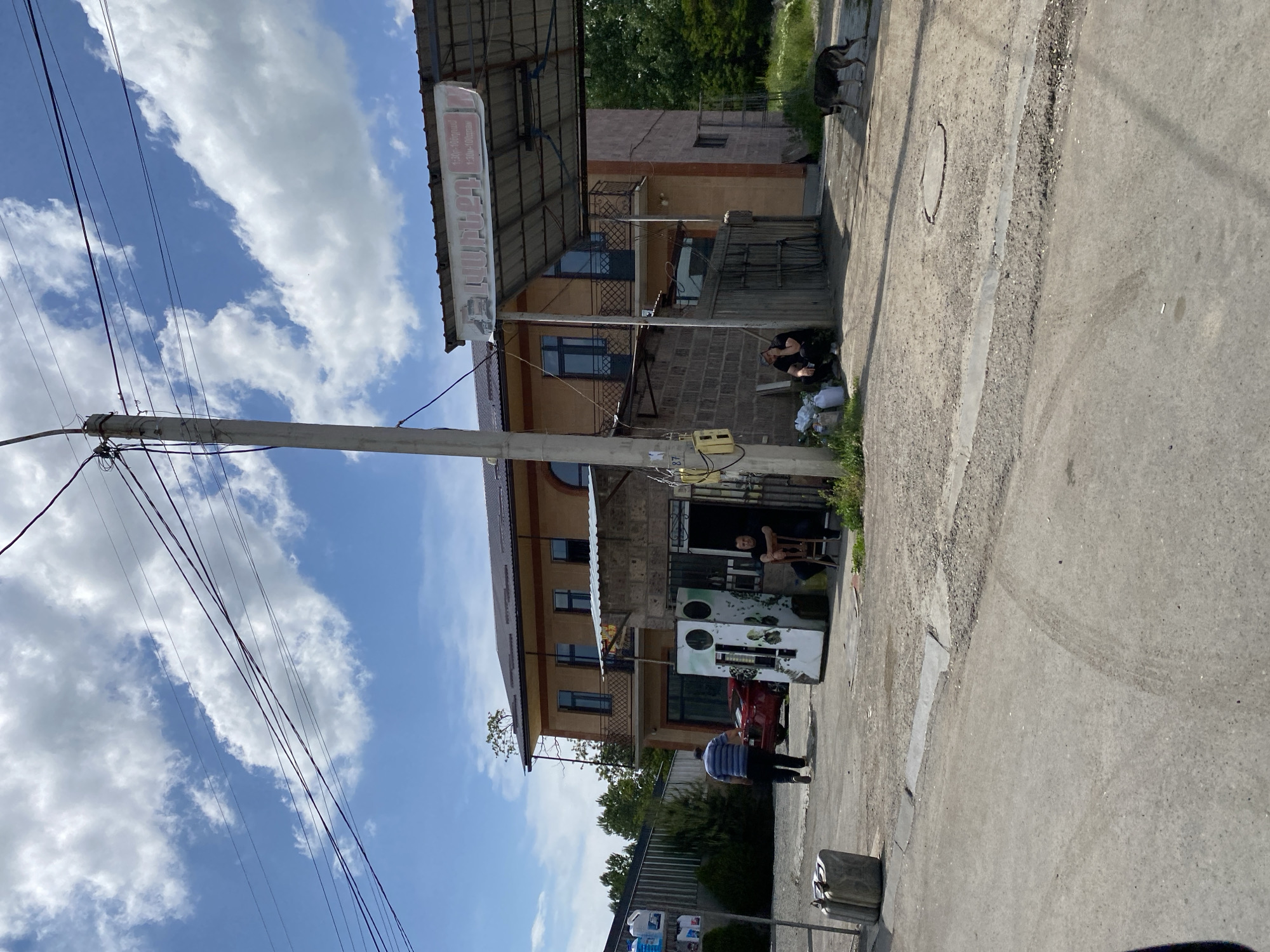 This media file depicts an outdoor scene on a sunny day, likely in Gyumri, Armenia. The image is oriented vertically. In the foreground, a paved area and a grassy verge are visible. A utility pole with numerous wires runs along the left side of the frame.
The middle ground features a two-story building with a rustic facade, constructed from what appears to be brick and stucco. A covered porch area extends from the ground floor, beneath which several objects are placed, including what looks like a water cooler or vending machine and a red car. Two individuals are present near the building's entrance; one is seated under the awning, and another, wearing a striped shirt, stands in front of the building.
Further back, another building with a darker roof and balconies is visible. Above the covered porch of the foreground building, a sign with text is mounted. A dark-colored dog is seen on the grassy area to the right of the buildings. The sky is a clear blue with scattered white clouds, suggesting pleasant weather. The presence of utility poles and wires indicates an urban or semi-urban setting. The overall impression is a casual, everyday scene in a community.