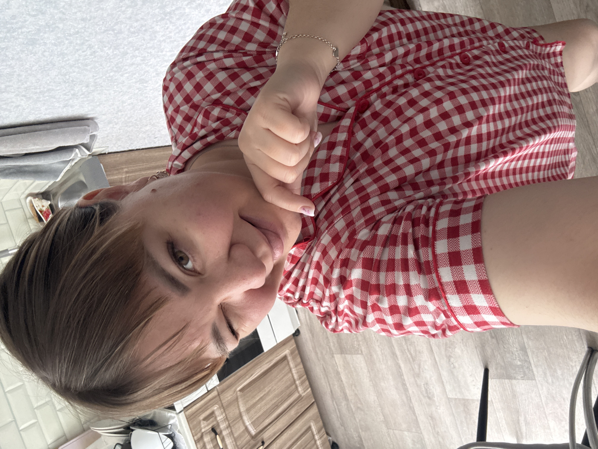 In this selfie, a woman with brown hair and bangs is captured from the chest up, leaning into the camera with a playful wink. She is wearing a red and white checkered shirt, and her right hand is raised to her chin, with her index finger pointing down. A thin silver bracelet adorns her left wrist. The background suggests an indoor setting, possibly a kitchen or living area, with wooden cabinets and a tiled wall visible. The floor appears to be light-colored wood laminate. The scene does not provide any specific indicators of its location within Stary Oskol, Russia, or any other city.