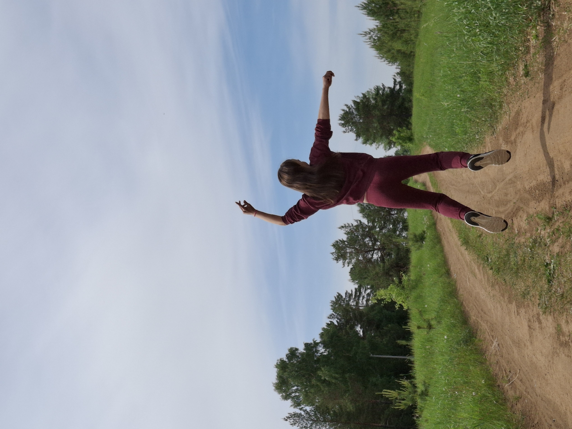 A photograph depicts a woman in a burgundy tracksuit and sneakers. She is positioned against a backdrop of blue sky with scattered clouds and green vegetation bordering a dirt path. The environment appears to be a natural area, with trees visible in the background.
Location details such as the city of Chaikovsky, Russia, are not visually verifiable from the provided image.