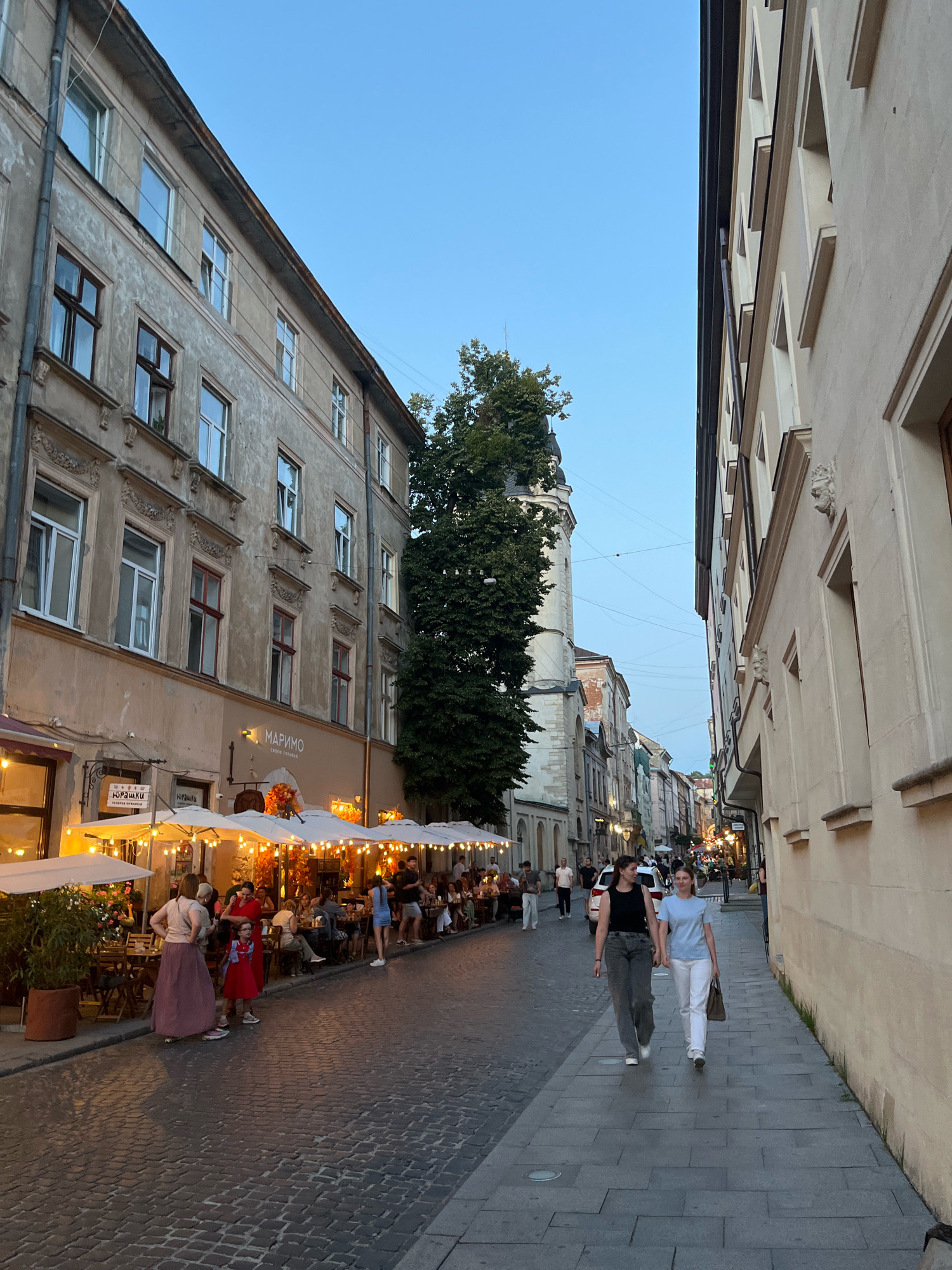 The image depicts a street scene in Lviv, Ukraine, likely during the early evening. On the left, a building with a weathered facade features multiple windows and balconies. A restaurant with outdoor seating is set up along the sidewalk, with white umbrellas providing shade over tables occupied by patrons. The cobblestone street is illuminated by warm lighting from the restaurant. Further down the street, other buildings and illuminated storefronts are visible.
On the right side of the street, a light-colored building with architectural details lines the sidewalk. Two women are walking hand-in-hand along this sidewalk, one in a black top and grey pants, the other in a blue t-shirt and white pants. The sky is a clear blue, indicating the twilight hour. The scene suggests a vibrant urban atmosphere with people enjoying an evening out.