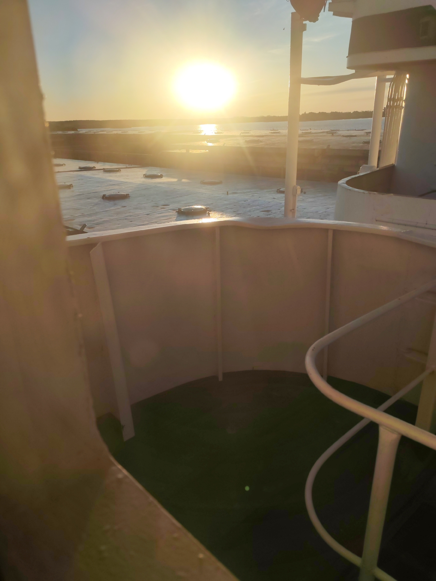 The image depicts a view from a structure, possibly a ship or building, overlooking a body of water with a bright, golden sun setting or rising on the horizon. The foreground features architectural elements like white railings and curved walls, suggesting an enclosed space with a green surface visible on the floor. The water in the background shows several dark, evenly spaced objects, possibly buoys or platforms. The light is intense, creating a strong contrast and casting long shadows.