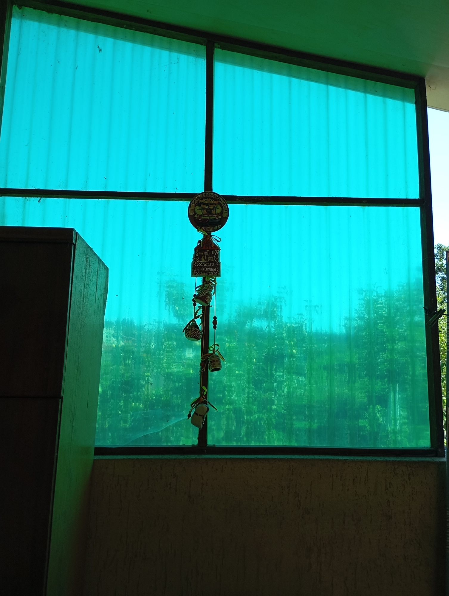 The image depicts a view through a turquoise corrugated plastic window, which is framed by a dark metal structure. Outside the window, lush green trees are visible, creating a blurred backdrop. Hanging from the window frame is a decorative wind chime. The wind chime is composed of several small, intricately designed ceramic or clay charms, including a round one at the top with what appears to be a painted scene, and a rectangular charm below it with inscribed text. Several miniature woven baskets and what look like tiny shoes are also suspended from the chime. To the left of the window, a dark, angular object, possibly a piece of furniture like a cabinet or wardrobe, is partially visible. The lower portion of the image shows a textured, light brown wall. The overall impression is a domestic or semi-outdoor setting, possibly a balcony or veranda, in a location that experiences significant greenery. The scene does not contain any people or animals.