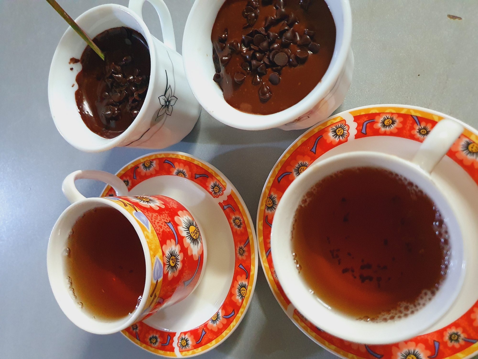 The image shows a close-up, overhead view of a breakfast scene with two cups of tea and two cups of chocolate pudding with chocolate chips. The cups are white with floral patterns. One cup of tea is on a saucer with the same floral pattern. The other cup of tea is on a separate saucer. The two cups of chocolate pudding are in plain white cups. A spoon is in one of the chocolate pudding cups. The setting appears to be a table. The lighting is natural and soft, suggesting it was taken during the daytime. The image is composed to showcase the food items, with a shallow depth of field that blurs the background. The location is not specified in the image or the prompt.