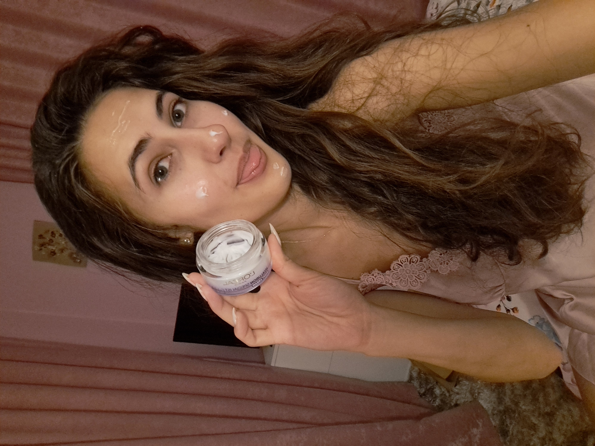 A young woman with long, wavy brown hair is holding a small jar of skincare cream and has dots of cream on her face, with her tongue sticking out. She is wearing a pink satin camisole. The background consists of pink curtains and what appears to be a dresser with a framed picture on it. The scene suggests a personal grooming or self-care moment.
