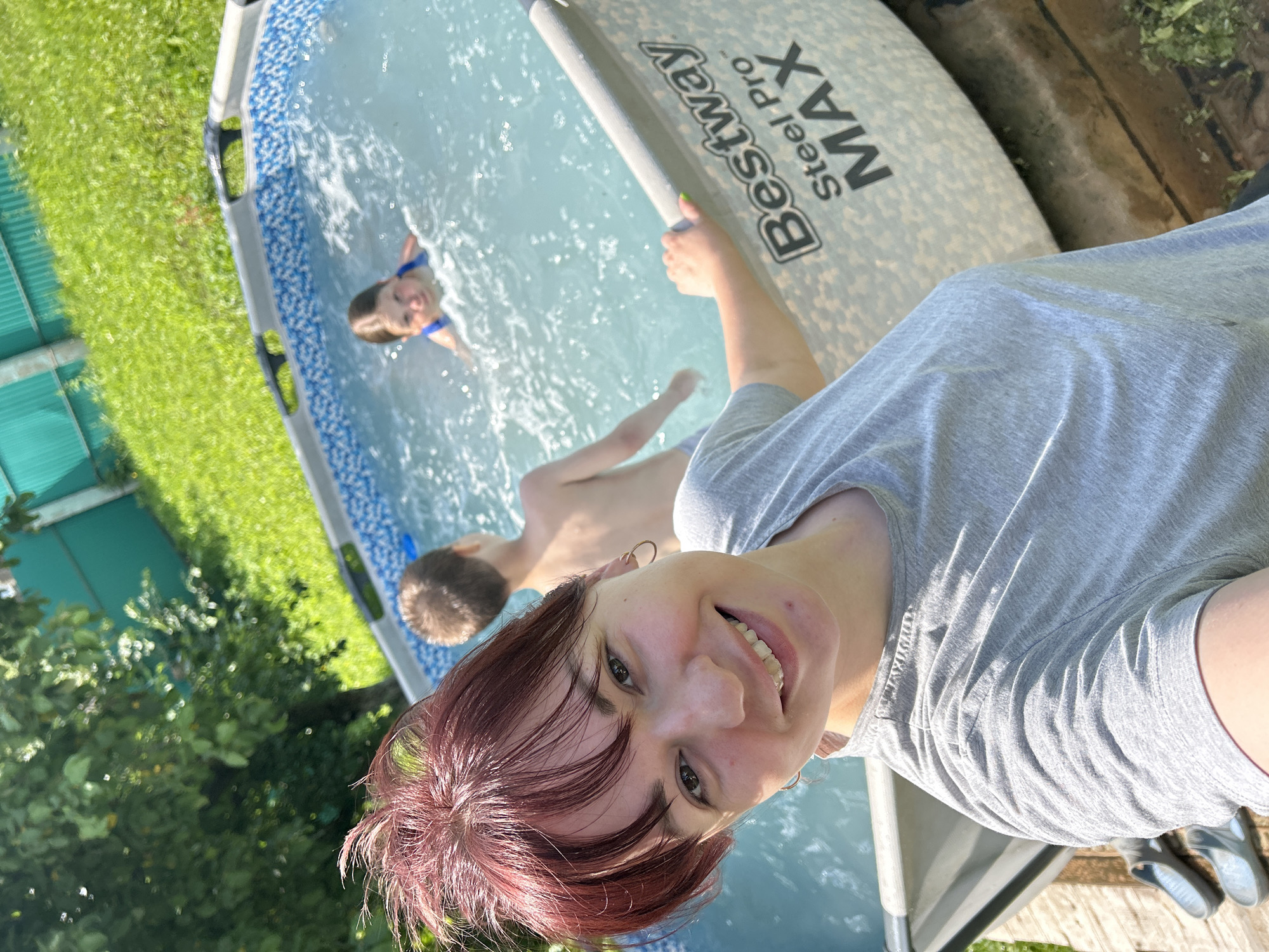 In a sunlit outdoor scene, a woman with reddish-brown hair pulled up in a bun smiles at the camera. She wears a gray t-shirt and is positioned in the foreground, close to a Bestway Steel Pro MAX swimming pool. Behind her, two children are in the pool; a boy is in the background, facing away, and a girl with dark hair, wearing a blue swimsuit, is closer to the foreground, splashing in the water. The pool has a blue and white patterned liner and is filled with clear water. The environment includes a green grassy lawn, lush green trees, and a teal fence in the background. Wooden planks are visible on the ground near the pool, with a pair of gray sandals placed on them. The overall setting suggests a casual, warm-weather activity, likely a backyard in the city of Foky, Russia.