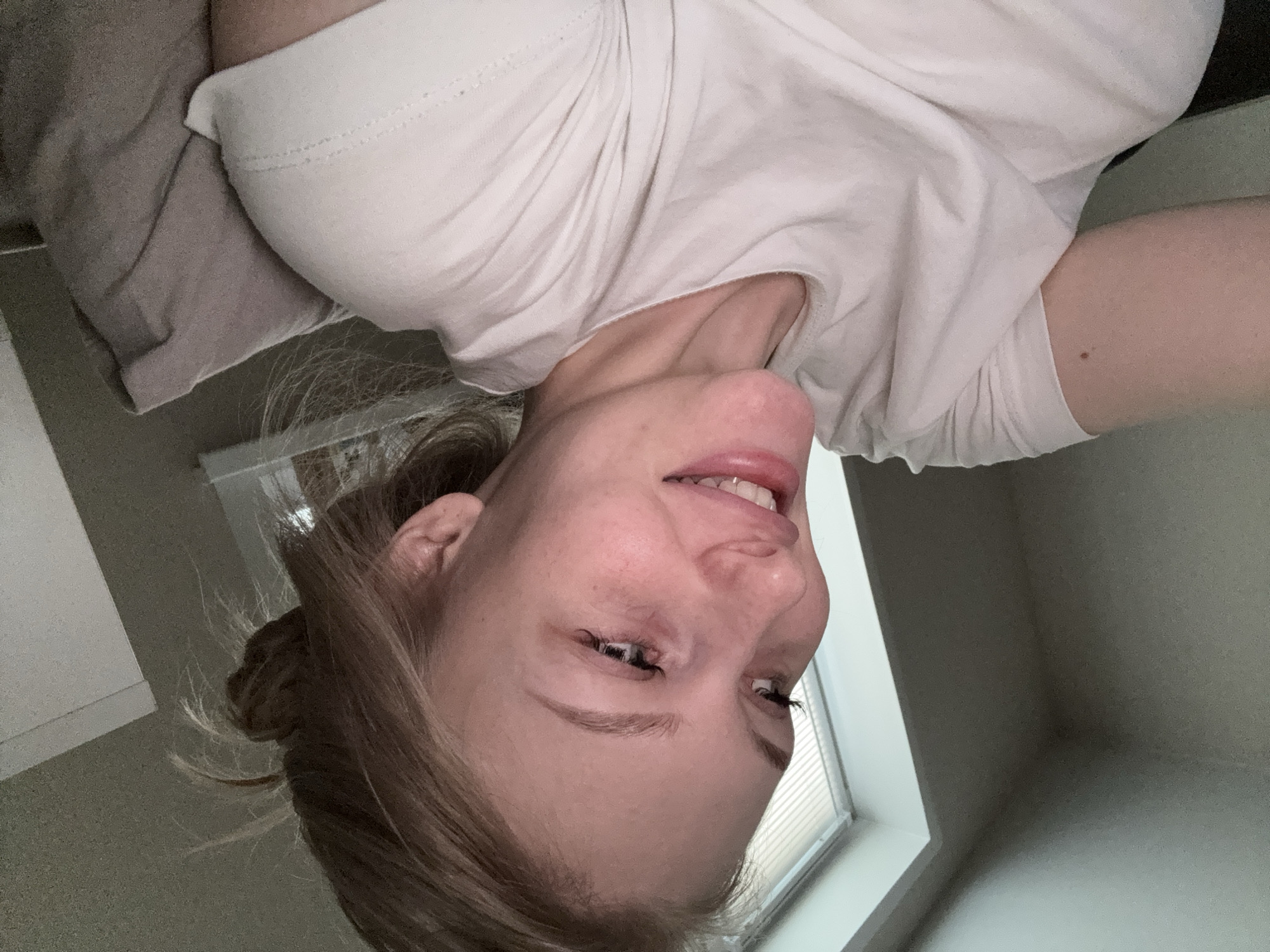 A young woman with blonde hair pulled back in a bun is captured in a close-up, self-taken photograph, looking to her left with a slight smile. She is wearing a plain white t-shirt and is positioned on a couch or bed, with a textured gray pillow visible behind her right shoulder. The background includes a window with blinds and a wall. The lighting suggests an indoor setting, possibly a home, with natural light coming from the window. The image is taken from the chest up. The overall scene is casual and domestic. The location is not specified within the image.