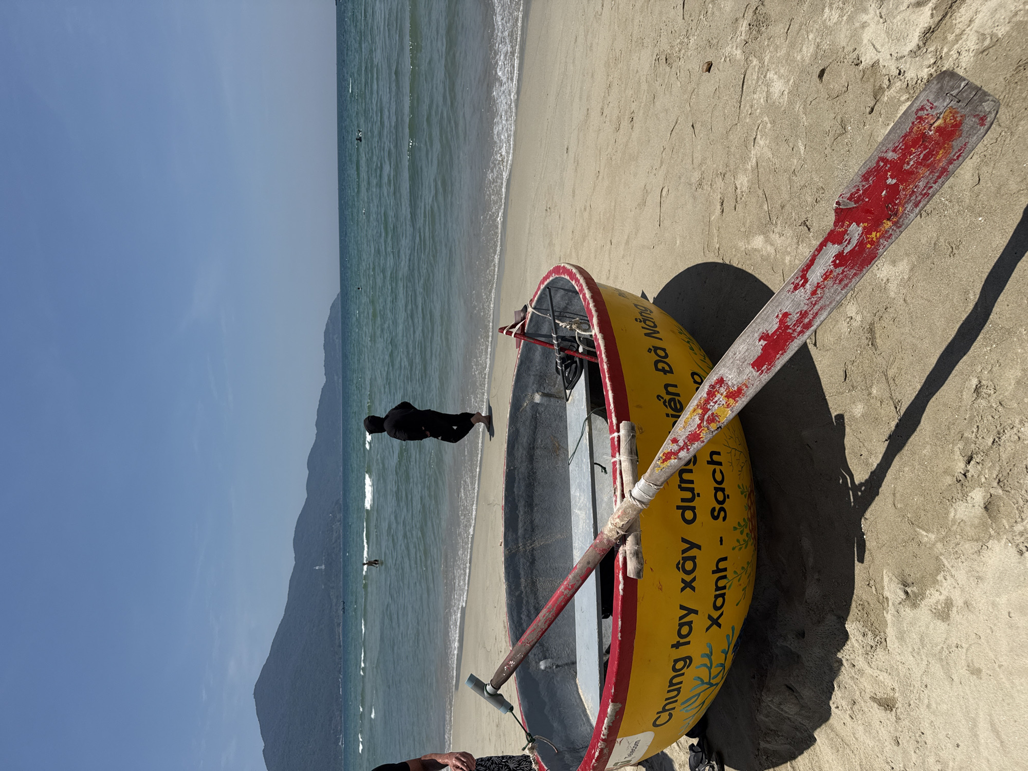 An eye-level, outdoor, daylight shot depicts a partially visible, overturned, yellow-and-red coracle on a sandy beach. The coracle has Vietnamese text. A worn, red-painted oar rests on top of it. The beach borders a sea with small waves and a dark silhouette of a person near the water's edge. In the background is a large, coastal mountain. The city is Da Nang, Vietnam.