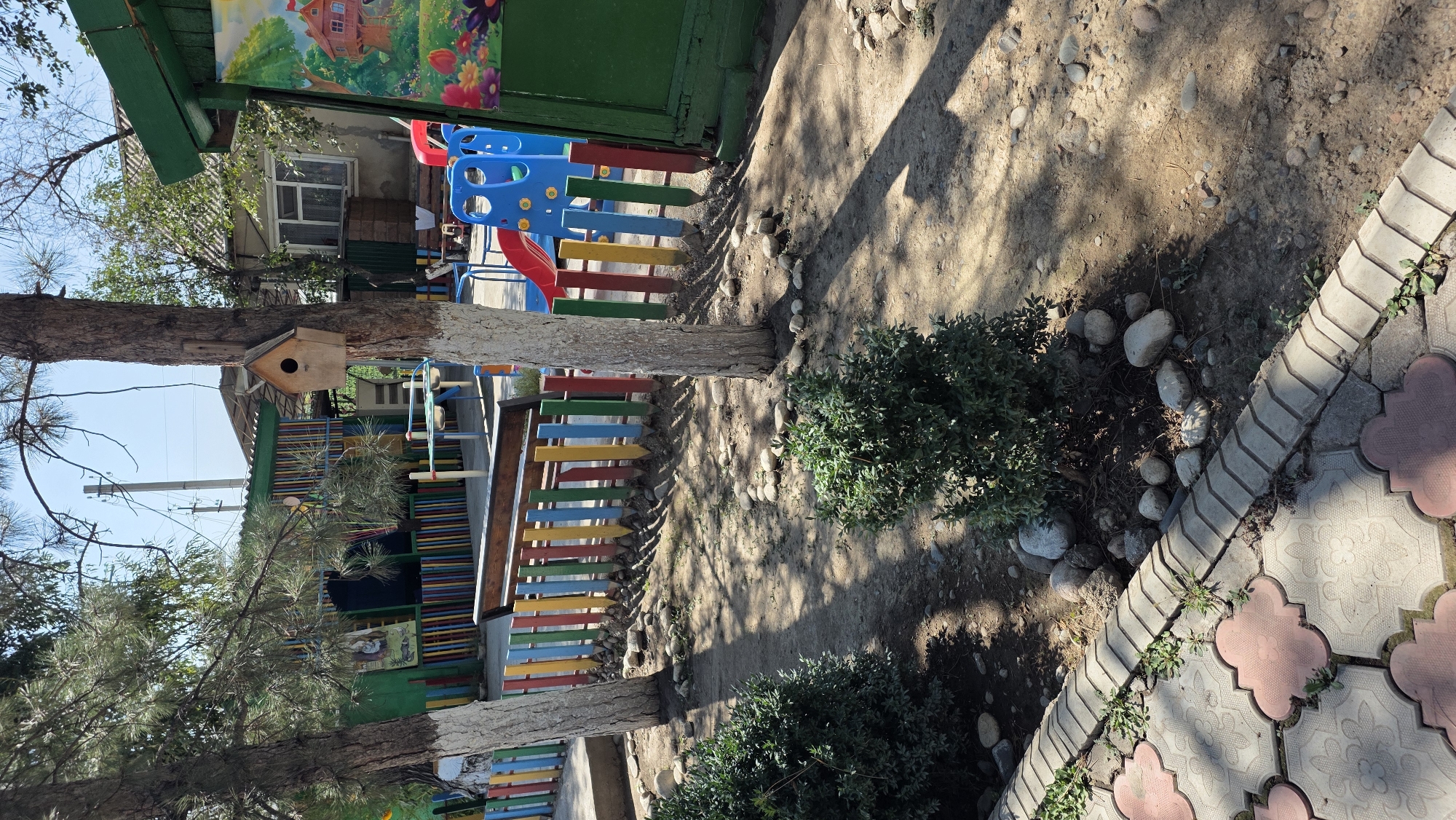 The image depicts a playground area with a colorful fence, a bench, a slide, and two trees. A wooden birdhouse is attached to one of the trees. The ground is covered in dirt and small stones. In the background, there is a building with windows and a painted mural on a green structure. The scene appears to be in a residential or communal area, possibly a kindergarten or park, located in Tokmok City, Kyrgyzstan. The weather is sunny, casting shadows on the ground. There are no people or animals visible in the image.