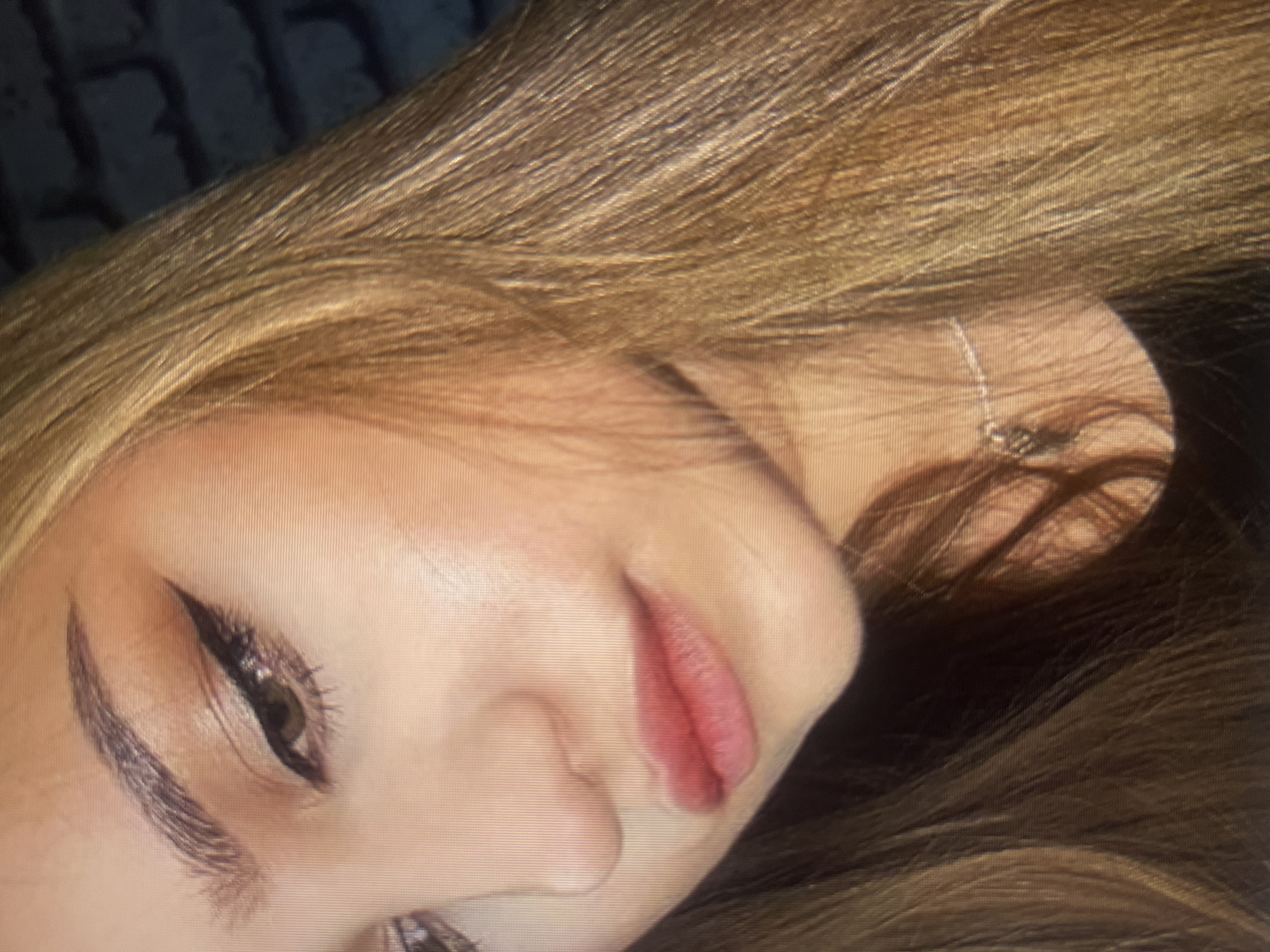 A close-up, low-angle medium shot shows a young woman's face from the nose up. She has light brown hair that falls across her face and shoulders. Her skin is fair, and she is wearing makeup, including eyeliner and lipstick. A silver necklace with a small pendant is visible around her neck. The background is blurred, showing a dark brick wall. The image appears to be taken indoors with artificial lighting. There is no discernible location in Qarağandı city administration, Kazakhstan.
