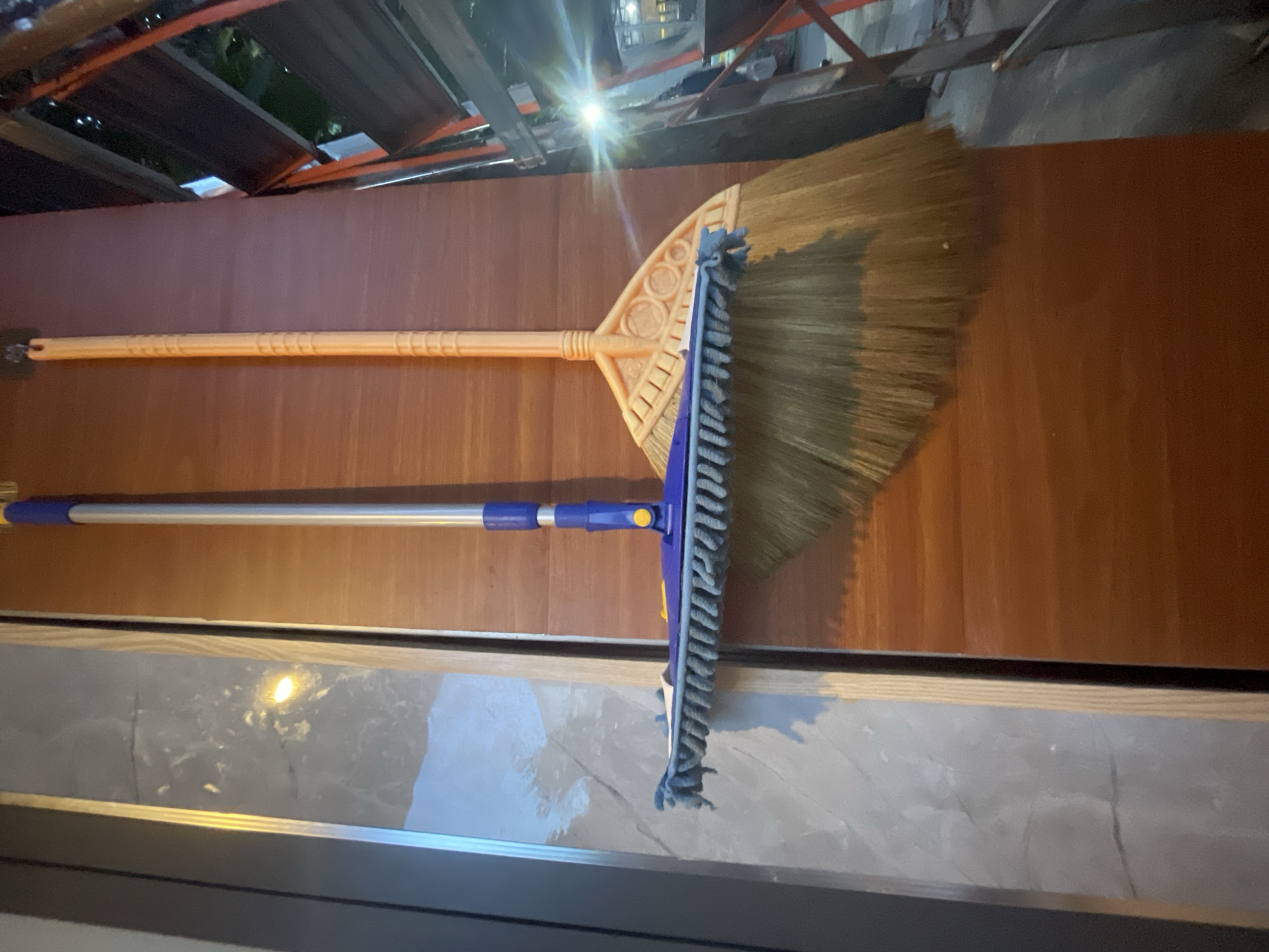 Two cleaning tools, a broom and a mop, are hanging vertically on a wooden wall. The broom, to the right, has a light orange handle and a fan-shaped head with straw bristles. The mop, to the left, has a silver pole with blue plastic accents and a rectangular head covered in blue microfiber strands. The background to the left of the wall appears to be a light-colored surface with a marbled or textured pattern. To the right of the wooden wall, a metal structure with visible planks and a bright light source is present, suggesting an outdoor or industrial setting. The lighting creates a glare on the right side of the image. The scene is static and offers no information about people or their actions. There is no contextual information available to determine the specific location in Phan Thiet Ward, Vietnam.