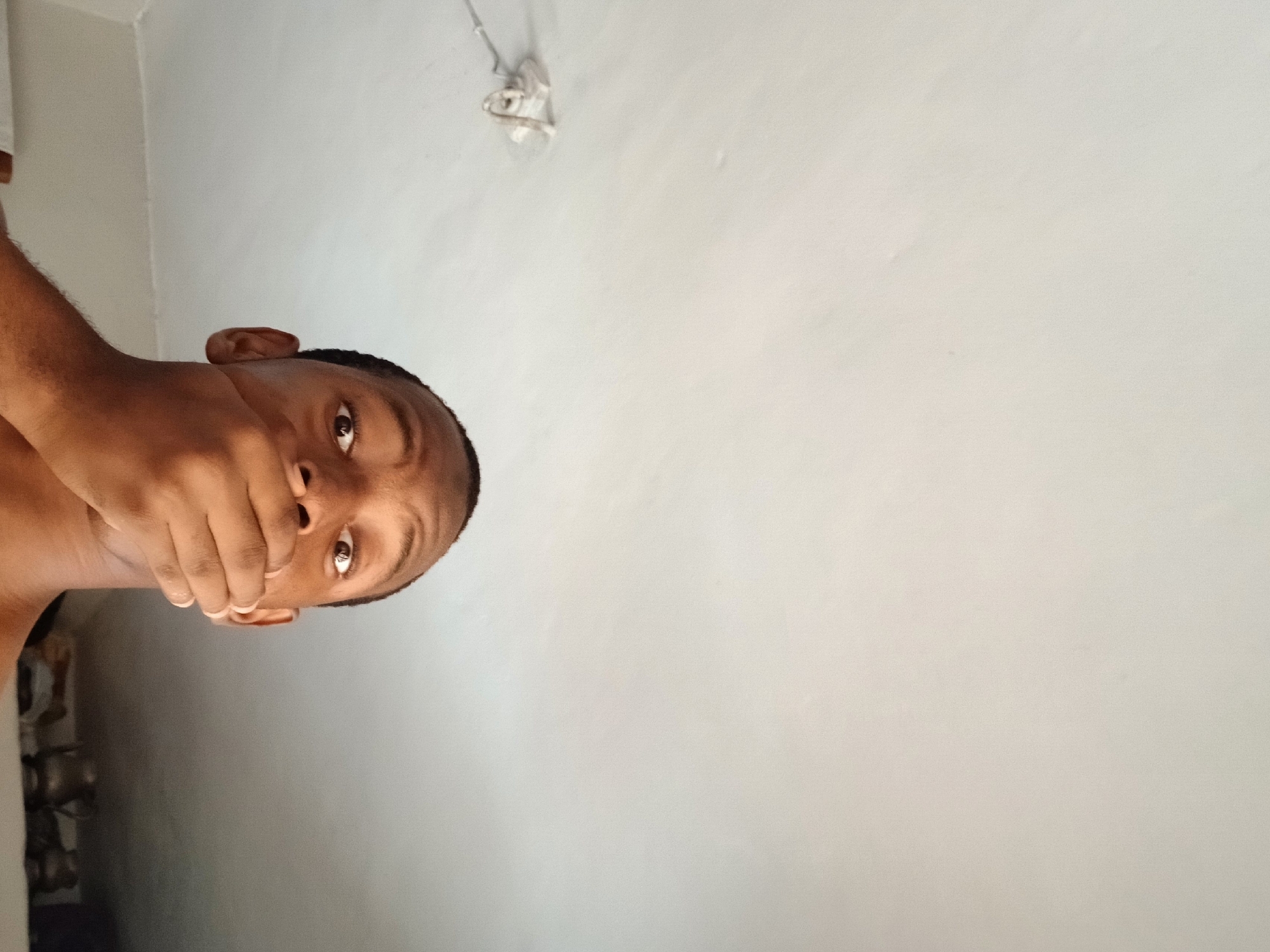 A dark-skinned man with close-cropped hair holds his hand over his mouth. He is looking directly at the camera. The background is a plain white wall with a light fixture near the top. The location is presumed to be Lagos, Nigeria, based on the prompt. No further location details are discernible from the image.