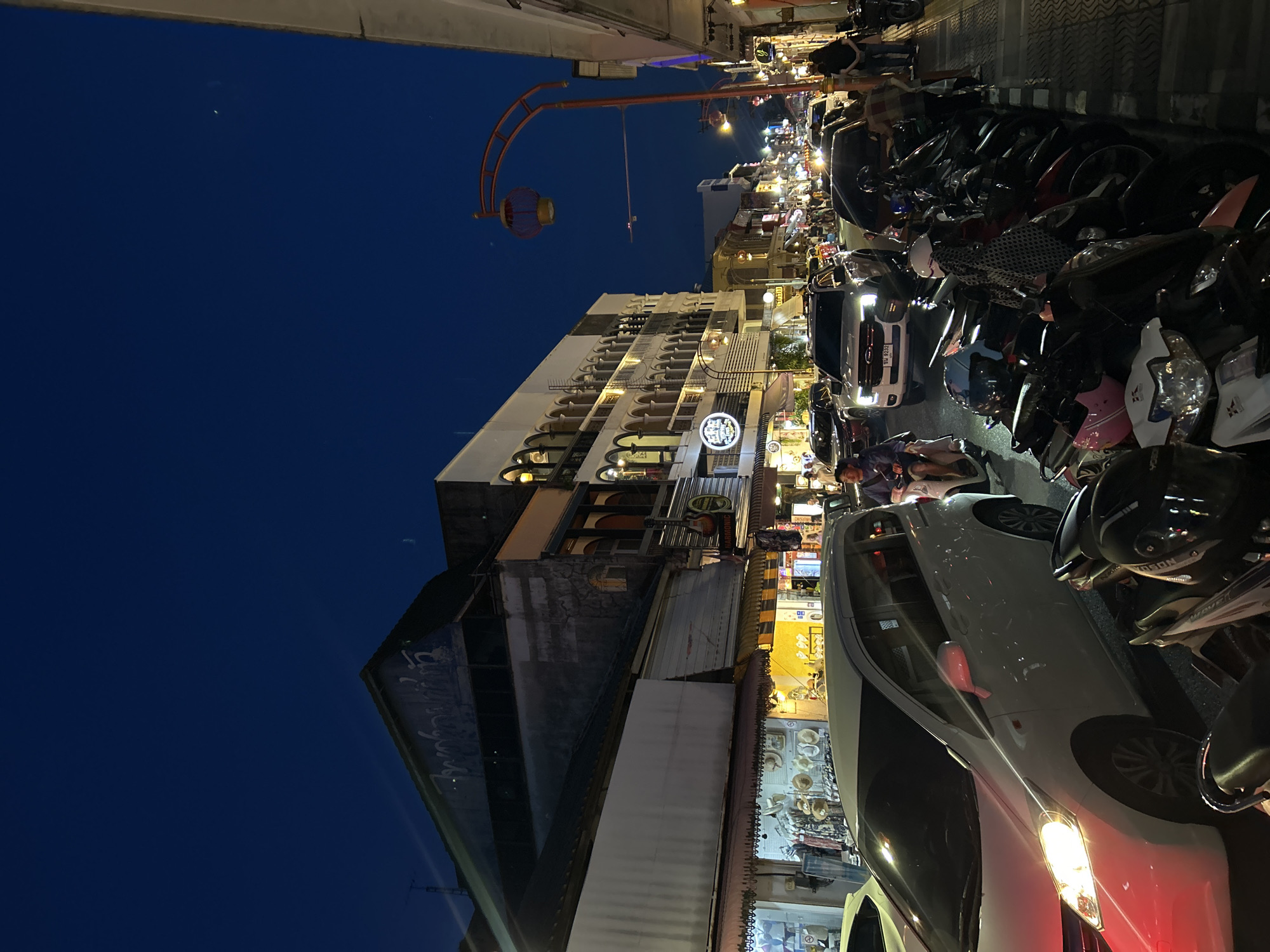 An outdoor, nighttime street scene in Phuket City Municipality, Thailand, shows a busy commercial street lined with shops and buildings. The sky is a deep, dark blue, with a faint crescent moon and stars visible. A street lamp with a red lantern hangs over the sidewalk.
The street is filled with a variety of vehicles, including cars and many parked motorcycles. A white minivan is prominent in the foreground, its headlights illuminated. A person is riding a scooter in the middle ground.
The buildings lining the street exhibit traditional architecture with arches and balconies. Some shops are illuminated, displaying merchandise such as clothing and hats. The overall atmosphere suggests a lively evening in a tourist area.