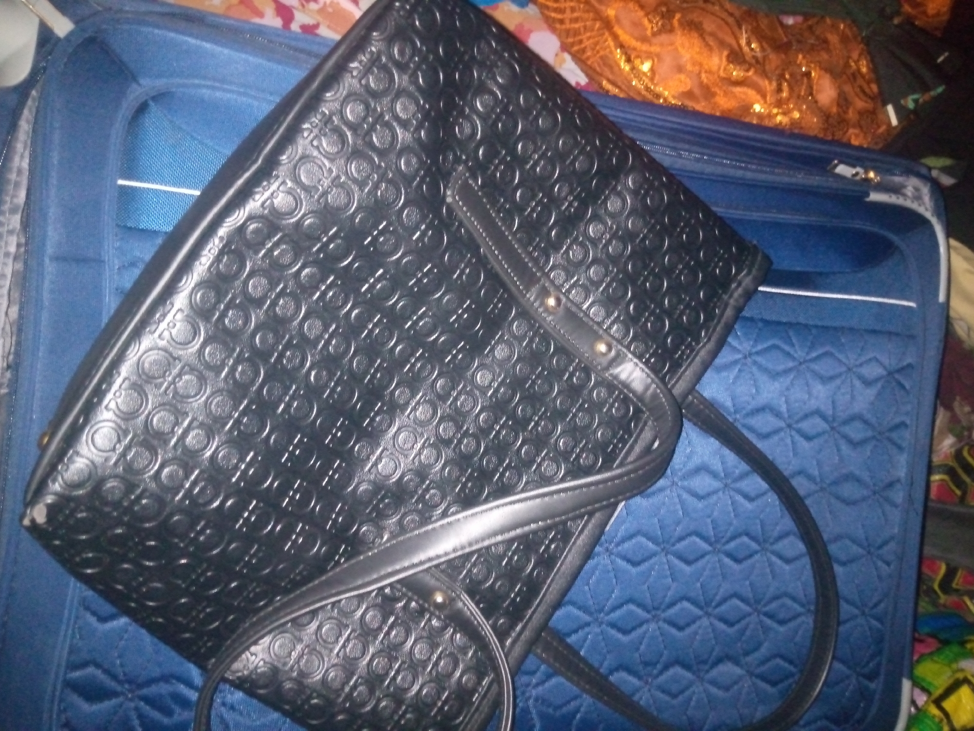 A black leather handbag with an embossed pattern of interlocking "G" logos is placed on top of a blue quilted suitcase. The handbag has two straps, one wider and one narrower, both made of black leather. The suitcase is partially visible and appears to be open. In the background, there is a piece of patterned fabric, possibly clothing or a blanket, in shades of orange and gold. The scene appears to be indoors, with the lighting suggesting it might be artificial. The location is unknown, in the city of Unknown, Unknown.