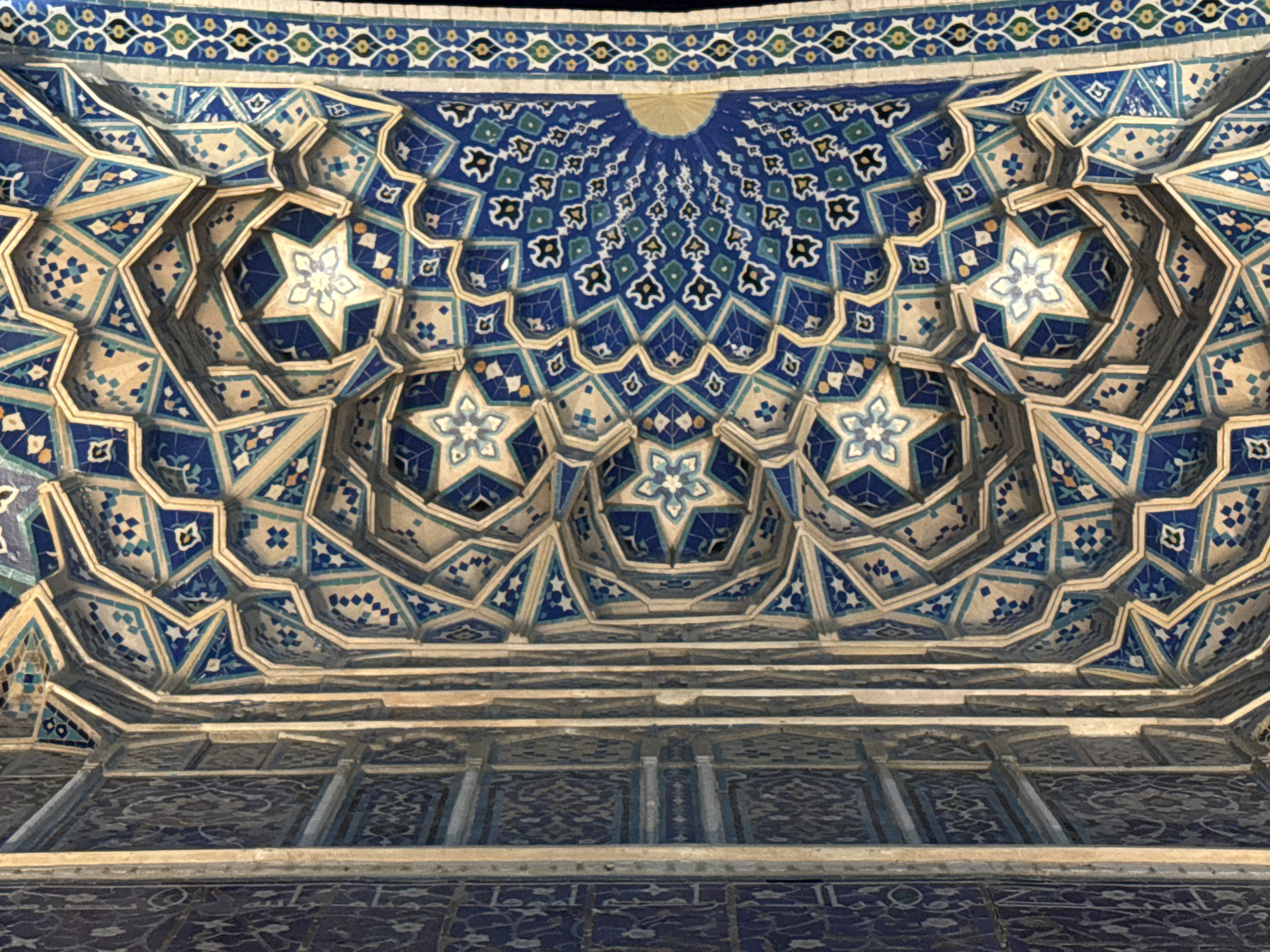 The image depicts the interior of a mosque or religious building in Samarkand City, Uzbekistan. The ceiling features intricate geometric patterns and star-shaped designs in shades of blue, white, and gold. The patterns are arranged in a repeating, symmetrical fashion, creating a visually complex and ornate surface. The lower portion of the image shows a wall or structural element with further patterned tiles and pillars. The overall effect is one of elaborate craftsmanship and traditional Islamic architecture.