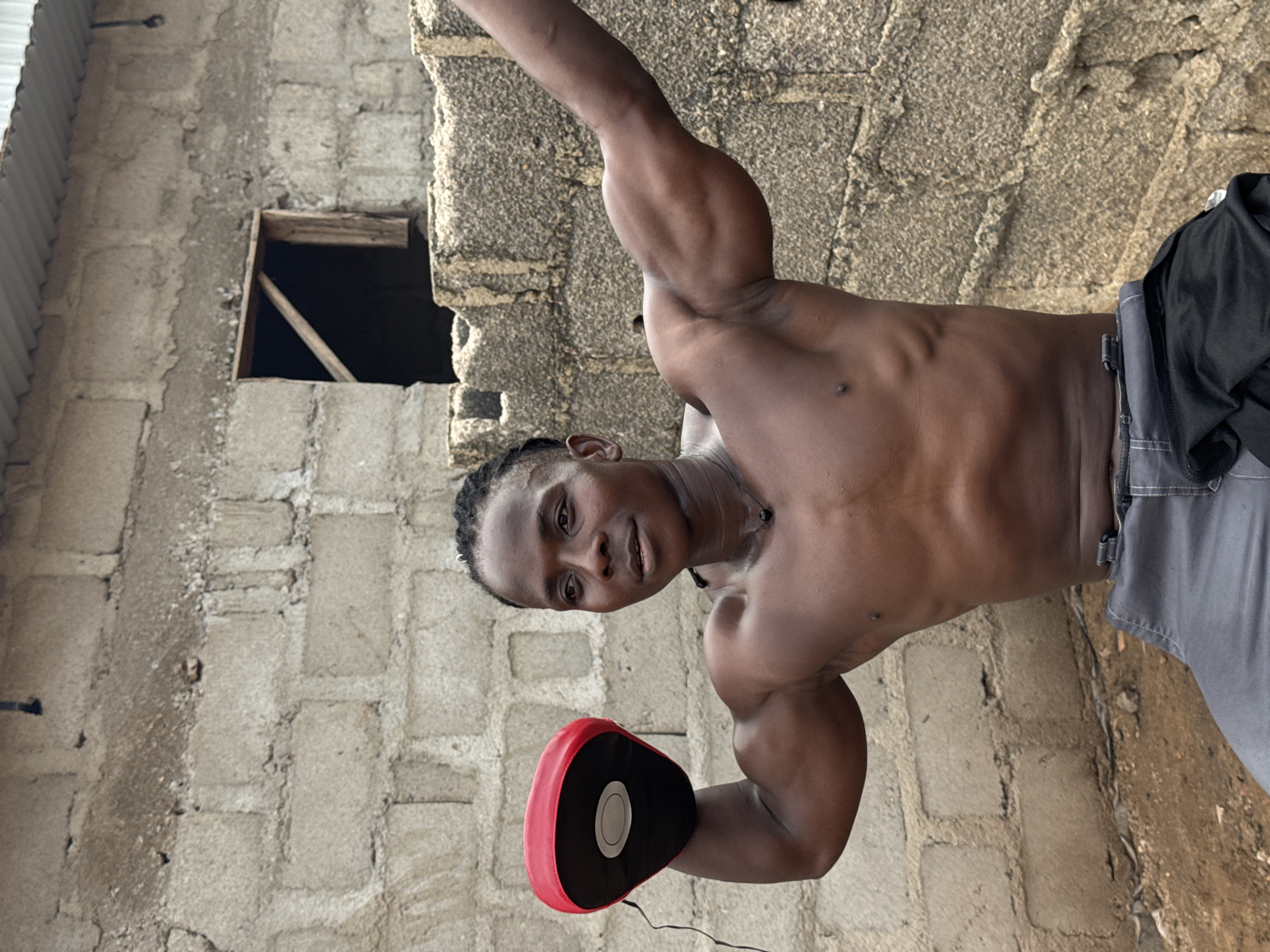 In Akwanga, Nigeria, a shirtless young man with dark skin and braided hair is posing in front of a weathered, rough-hewn brick wall. He is holding a red and black boxing pad in his left hand, which is raised. His right arm is extended upwards and slightly back, showcasing his muscular bicep. The man is wearing grey trousers. The environment appears to be an outdoor, somewhat dilapidated setting, with a corrugated metal roof visible in the upper right corner and a dark opening in the brick wall behind him. The ground is covered in dirt and small stones.