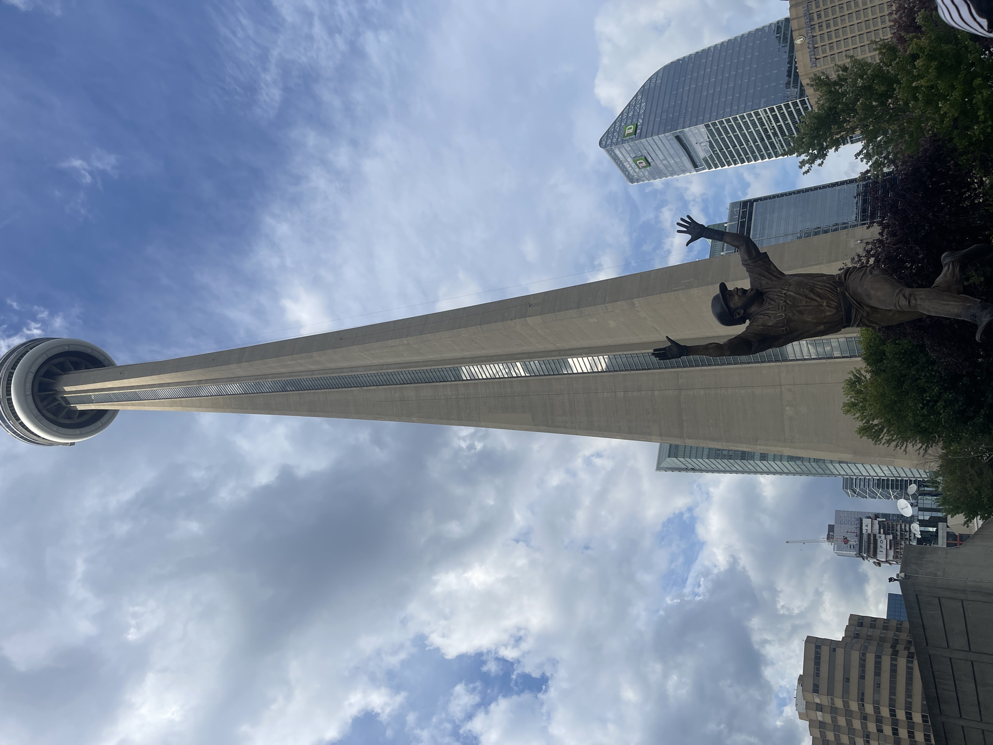 This image depicts the CN Tower in Toronto, Canada, as viewed from below. In the foreground, a bronze statue of a baseball player in mid-action, with arms raised, is visible. The statue appears to be a memorial. Behind the statue, the imposing concrete structure of the CN Tower rises into a cloudy sky. To the right of the CN Tower, a modern skyscraper with a distinctive curved design and the TD Bank logo is prominent. Other buildings and trees are also present in the background, contributing to the urban cityscape. The sky is a mix of blue patches and grey clouds, suggesting variable weather conditions.