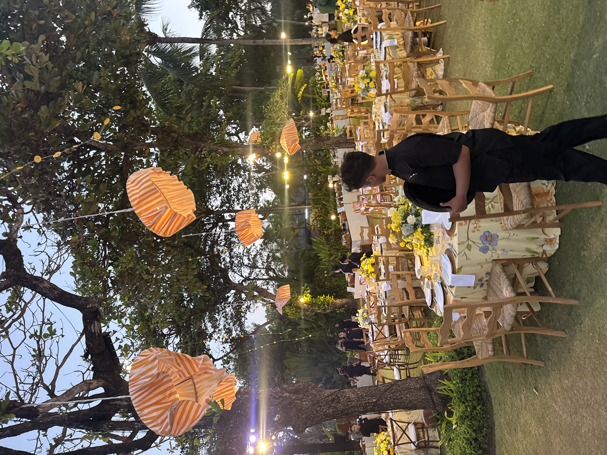 This image captures an outdoor event setup in Kuta Selatan, Indonesia, likely a wedding reception or a formal dinner. The scene is set in a garden area with lush green grass and large trees adorned with orange and white striped lanterns, adding a festive and whimsical touch. String lights are also visible, suggesting the event is taking place in the late afternoon or early evening.
In the foreground, a young man dressed in a black uniform is walking, carrying a black tray. He appears to be a server or staff member. The tables are covered with floral patterned tablecloths in yellow and blue hues, and they are meticulously set with plates, napkins, and glassware. Centerpieces of yellow and blue flowers enhance the decor.
In the background, more tables and chairs are arranged, and several people, also dressed in dark uniforms, are seen attending to the setup or serving guests. Further back, buffets are set up with food. The overall impression is one of an organized and elegantly decorated outdoor gathering. The location appears to be a resort or a private venue in Kuta Selatan, known for its beaches and tourist accommodations.