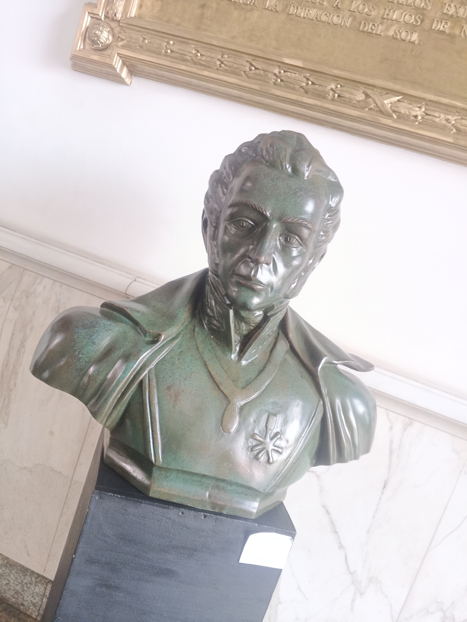 The image shows a bronze bust of a man in military attire displayed indoors. The bust is set atop a dark gray pedestal. The background includes a white wall with marble-like veining and a framed plaque with gold trim visible above and behind the bust. The plaque contains text in a language other than English, mentioning "LA DURACION DEL SOL." The bust's features are detailed, suggesting a historical or commemorative piece. The setting appears to be a museum or government building in an unknown city, as noted in the prompt.
